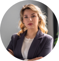 Portrait of a woman with blonde hair wearing a dark blazer and white top, standing against a neutral background.