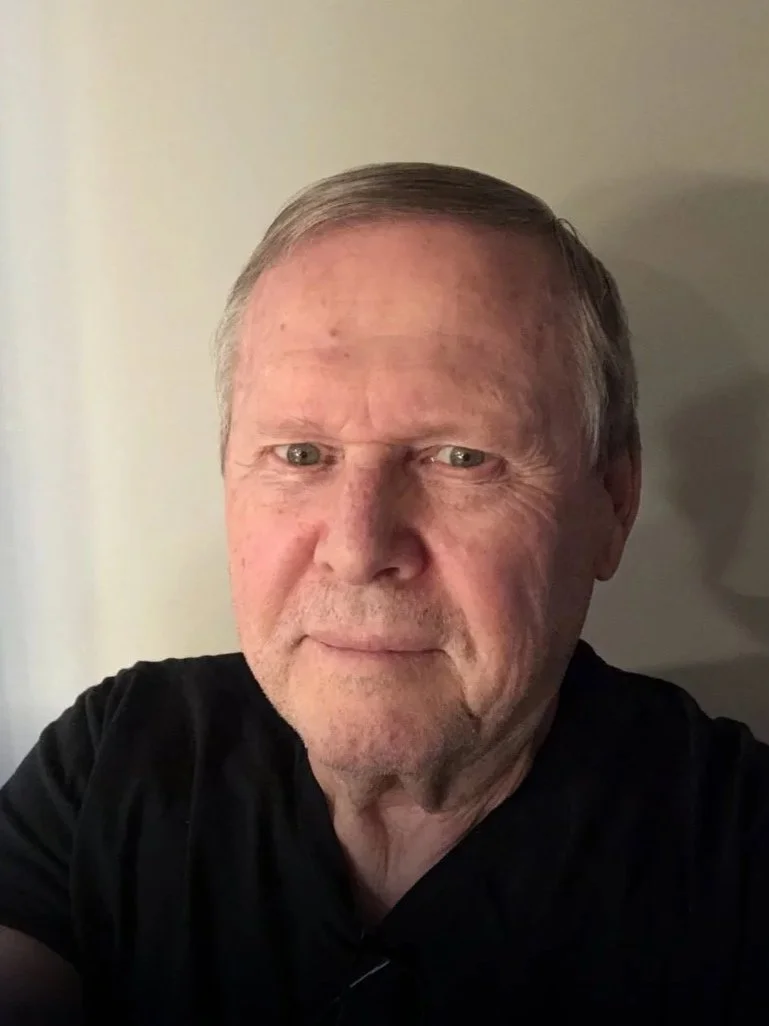 A close-up selfie of an older man with fair skin, short gray hair, and blue eyes, wearing a black shirt, indoors in front of a plain light-colored wall.