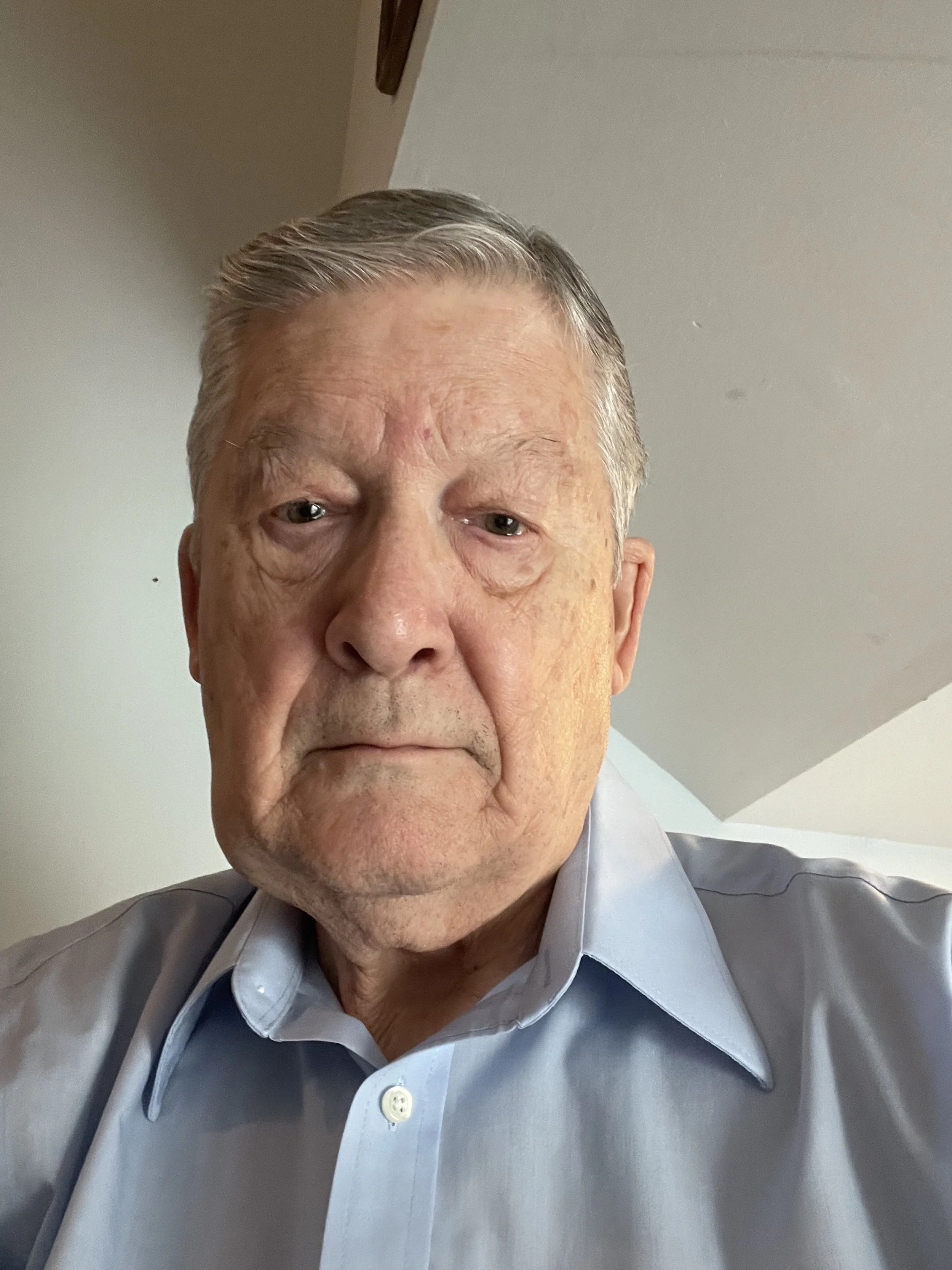 A close-up photo of an elderly man with short gray hair, wearing a light blue collared shirt, looking directly at the camera with a neutral expression. The background shows a part of a ceiling and wall.