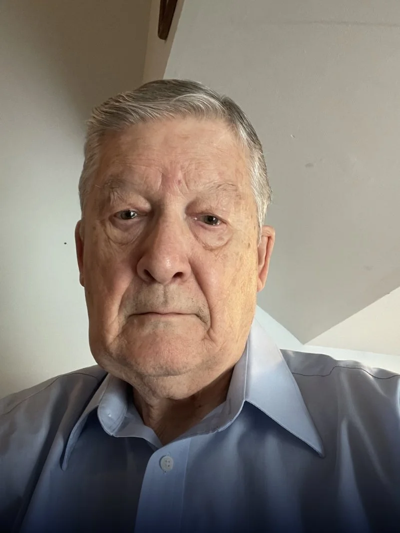 A close-up of an elderly man with short gray hair, wearing a light blue collared shirt, in a room with a sloped ceiling.