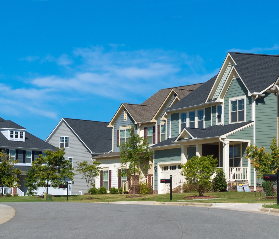 A neighborhood street with multiple colorful two-story houses, green lawns, trees, and mailboxes under a bright blue sky.