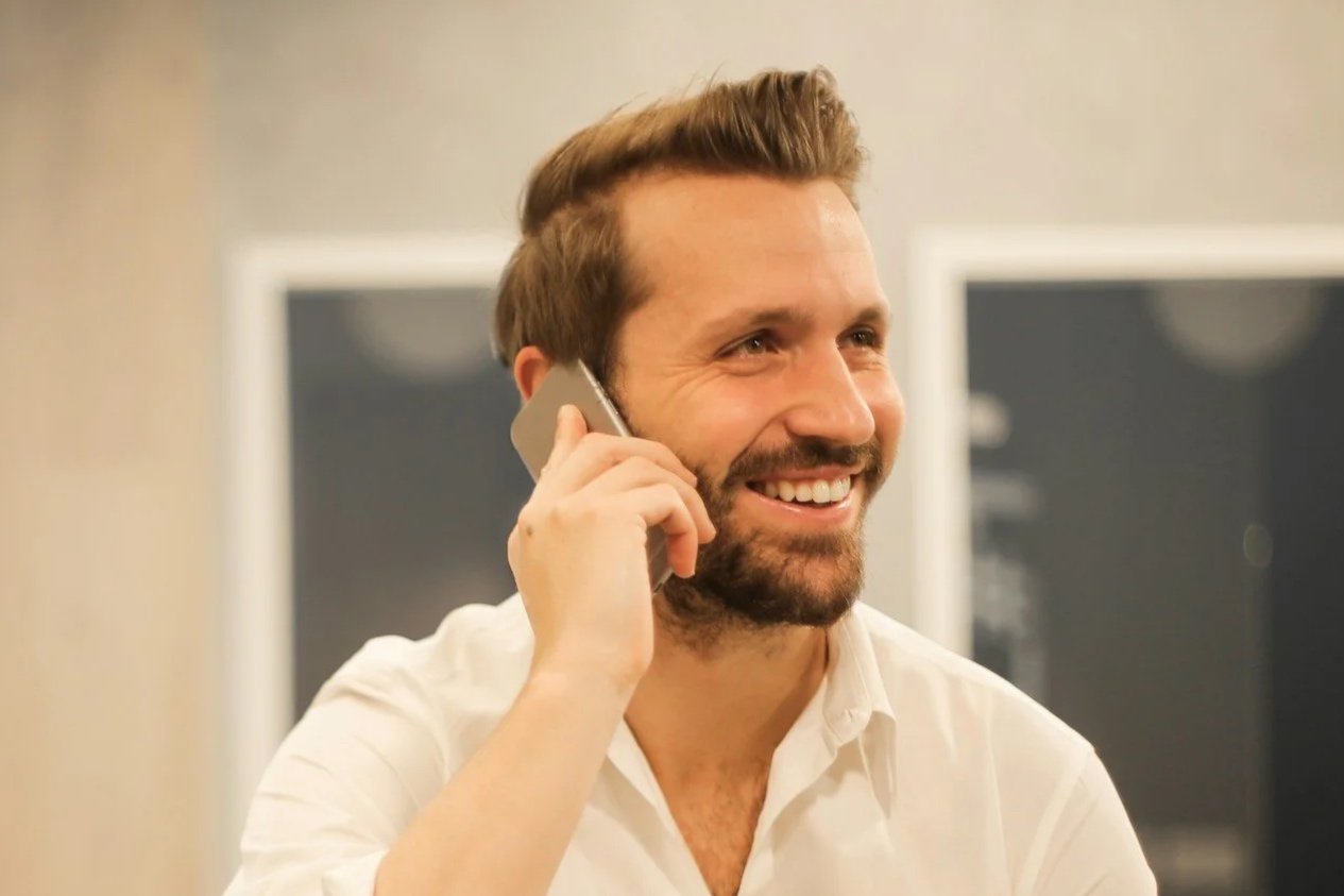 A man with red hair and fair skin wearing a navy blue suit and white shirt, sitting at a desk with a computer monitor.