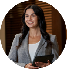 A woman with long dark hair, wearing a gray blazer over a white top, is smiling and holding a tablet in her right hand.