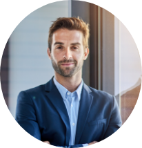 A man in a blue suit smiling and standing indoors near a window.