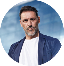 A man with short dark hair and a beard, wearing a navy blazer and a white shirt, standing outdoors with a cloudy sky background.