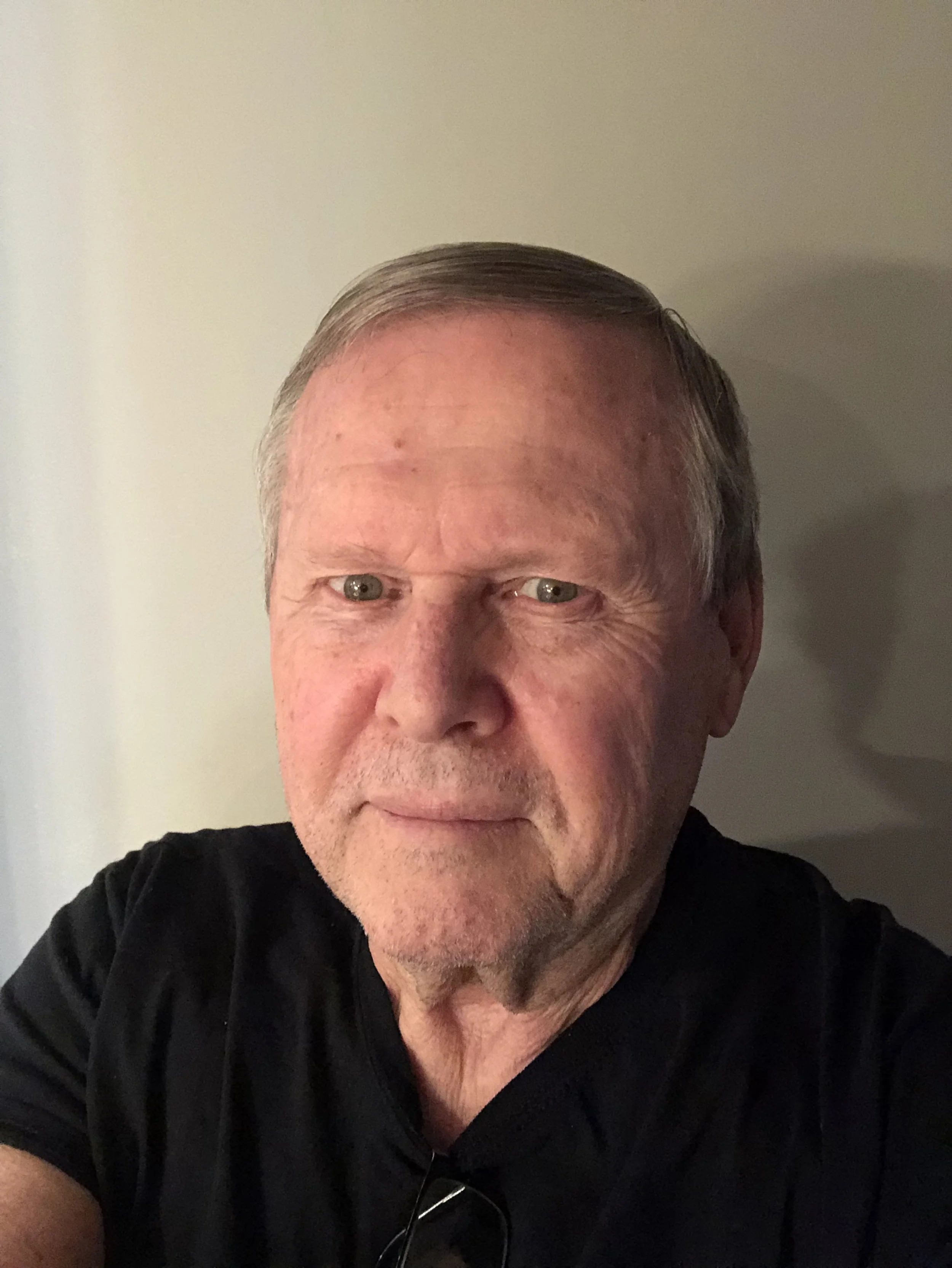 A middle-aged man with short gray hair, blue eyes, and a light beard, wearing a black shirt, taking a selfie in a room with plain light-colored walls.