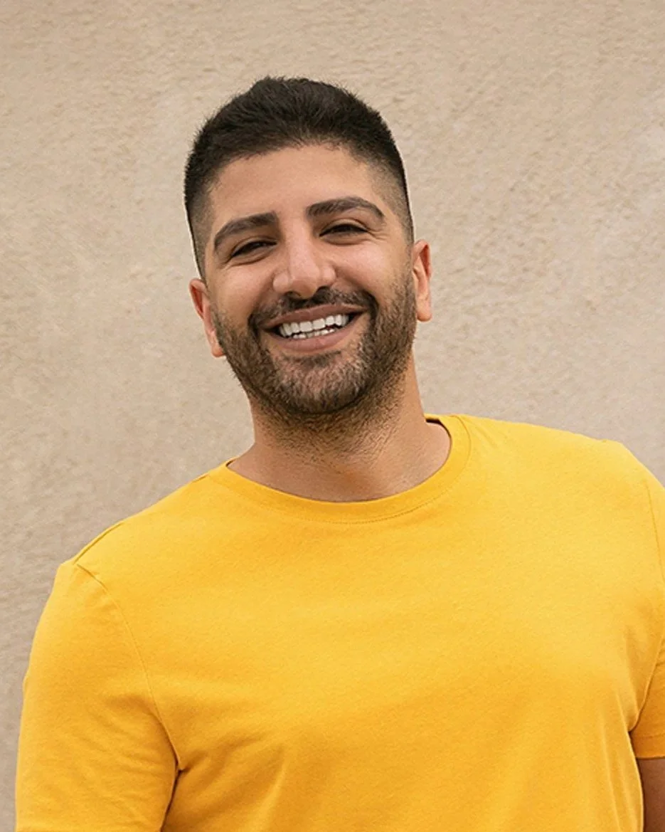 A man with dark hair and a beard wearing a dark suit, posing with his hand on his chin outdoors with green foliage in the background.
