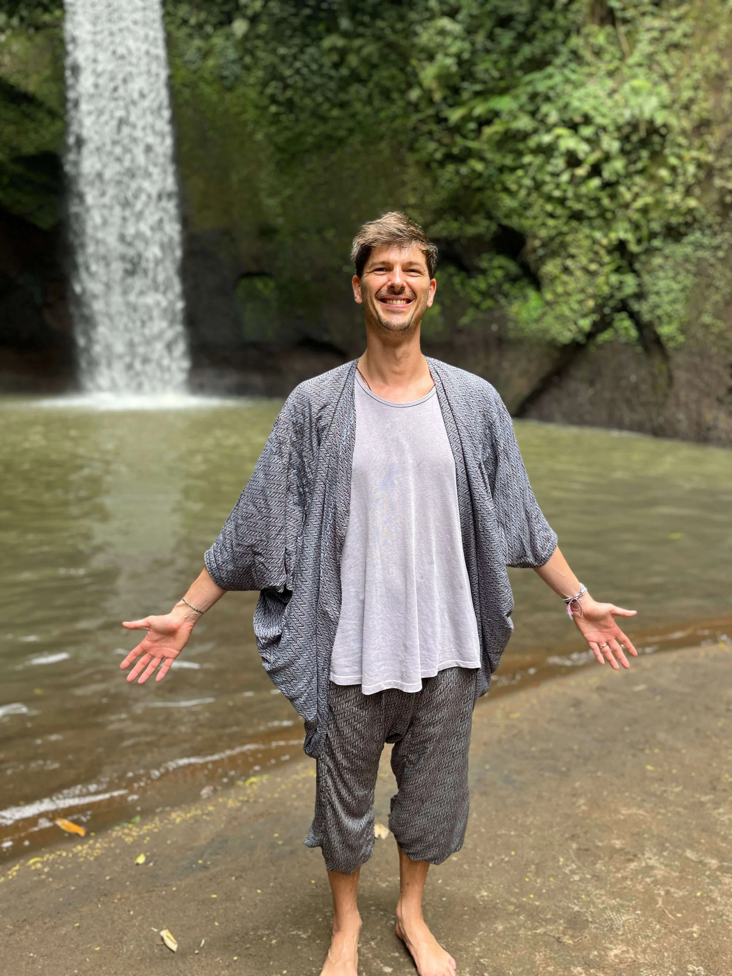 Mathis standing barefoot in water near waterfall, smiling and extending arms, surrounded by lush green foliage. A community to find career fulfillment in tech, offered by Coach Mathis Lauckner to help tech professionals navigate crossroads.