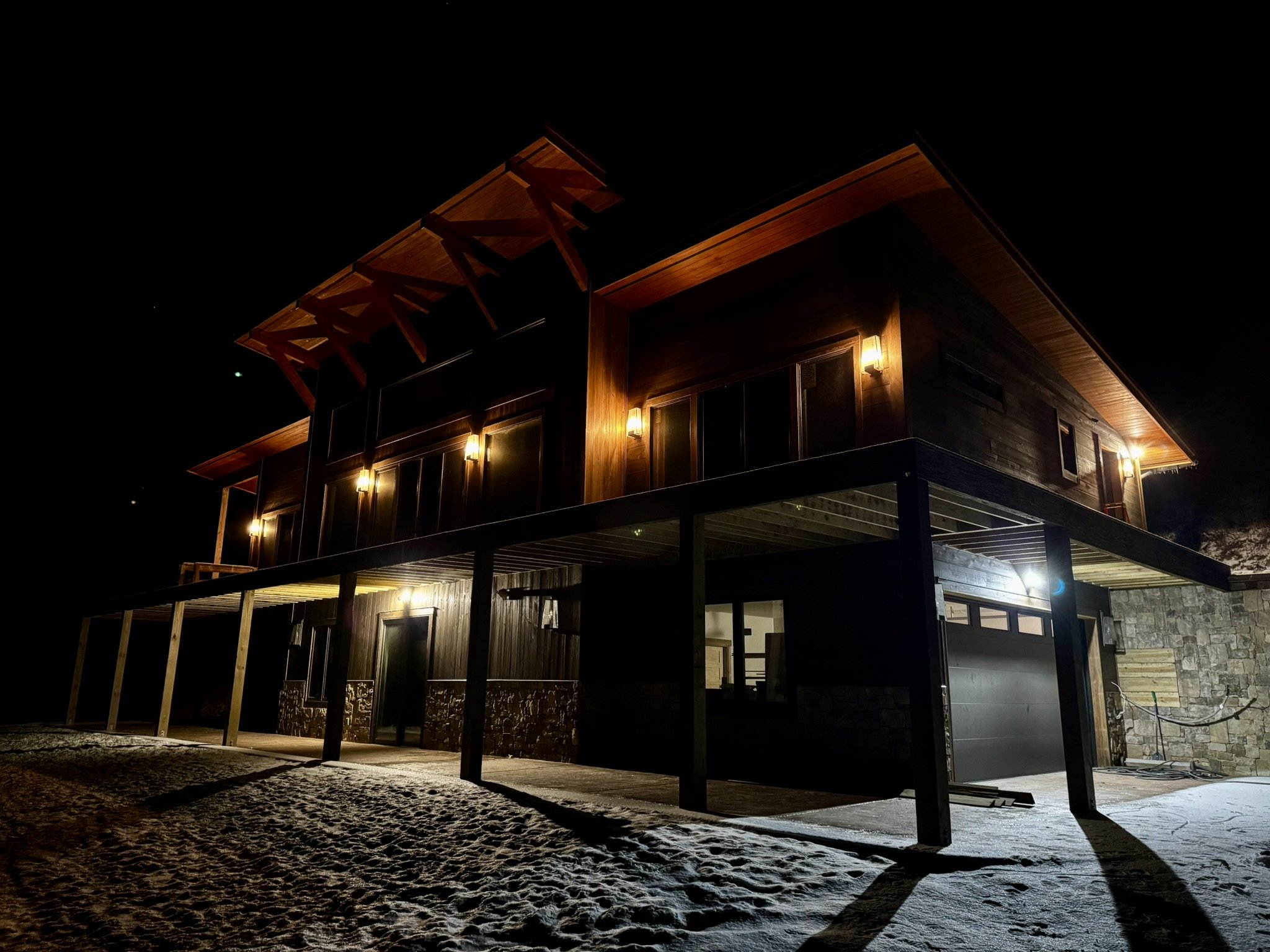 Night view of a modern two-story house with wooden exterior, lit by exterior lights, and snow on the ground.