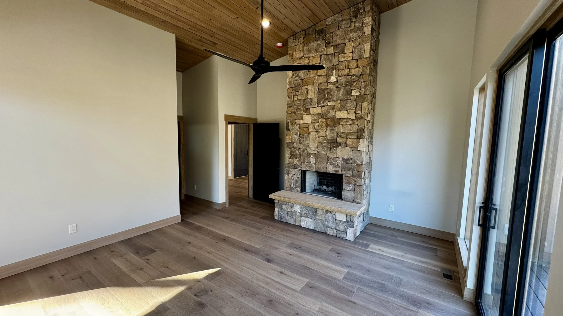 Living room with wooden floors, a stone fireplace, a ceiling fan, and sliding glass doors.