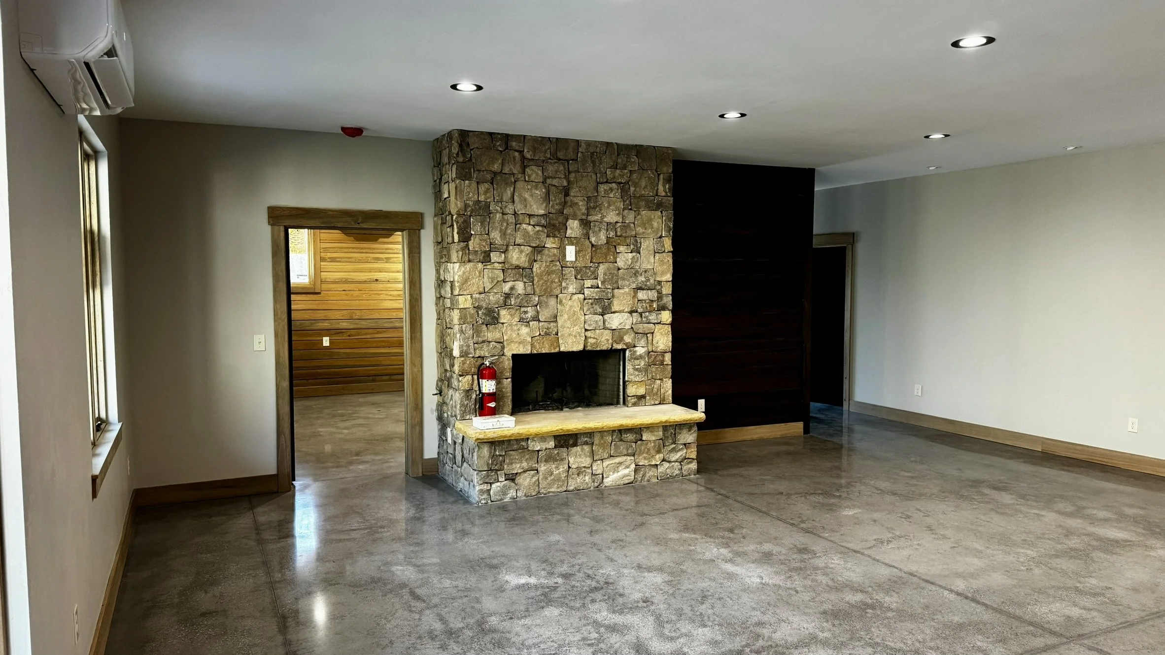 Empty living room with a stone fireplace, concrete floor, and beige walls. There is a fire extinguisher on a ledge next to the fireplace, and a doorway leading to a room with wood-paneled walls. Recessed ceiling lights are installed.