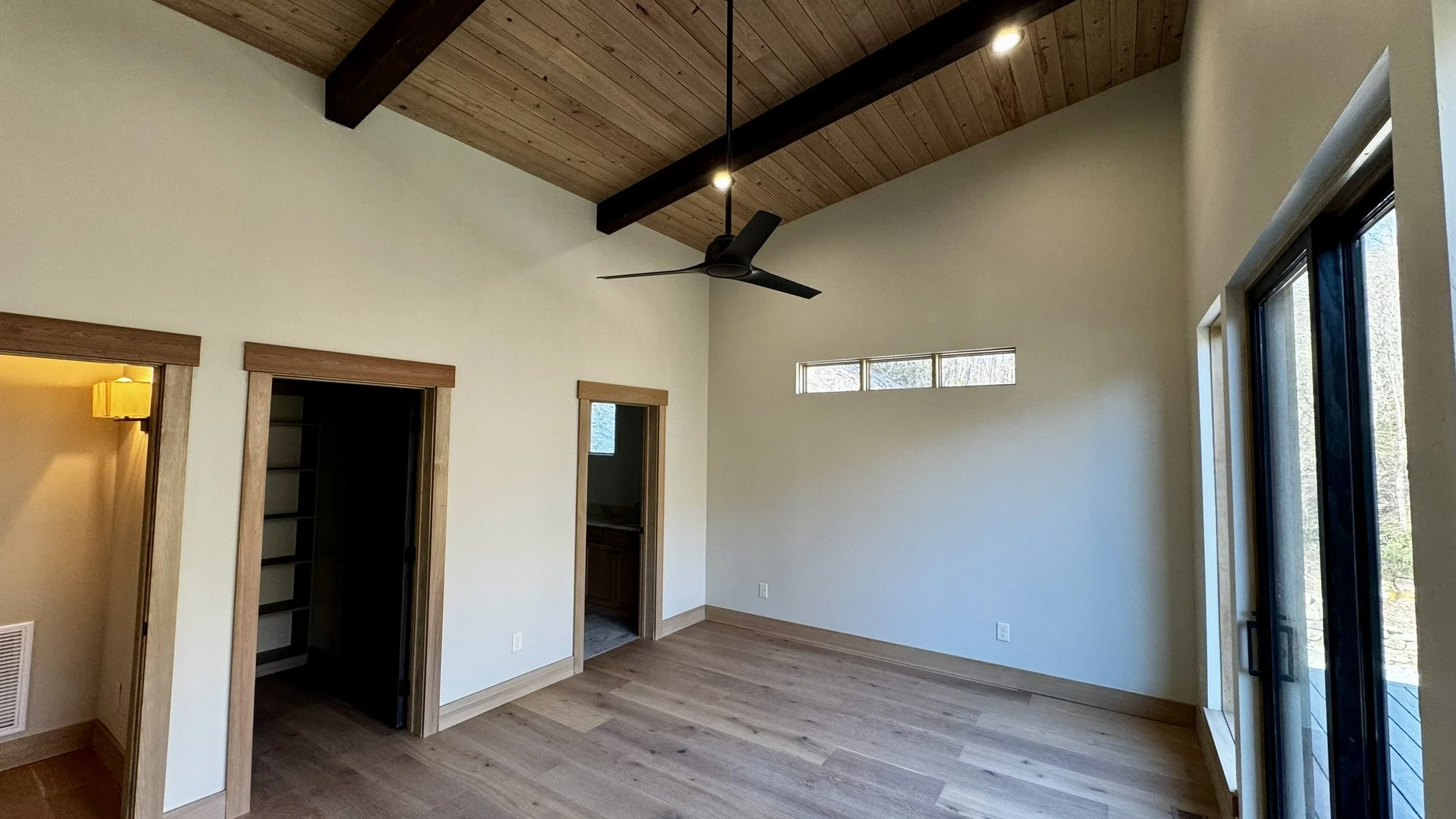 Empty room with wooden floors, beige walls, a high ceiling with wooden panels and dark beams. Ceiling fan, small horizontal window, and sliding glass door leading outside. Two doorways showing closet space and a bathroom.