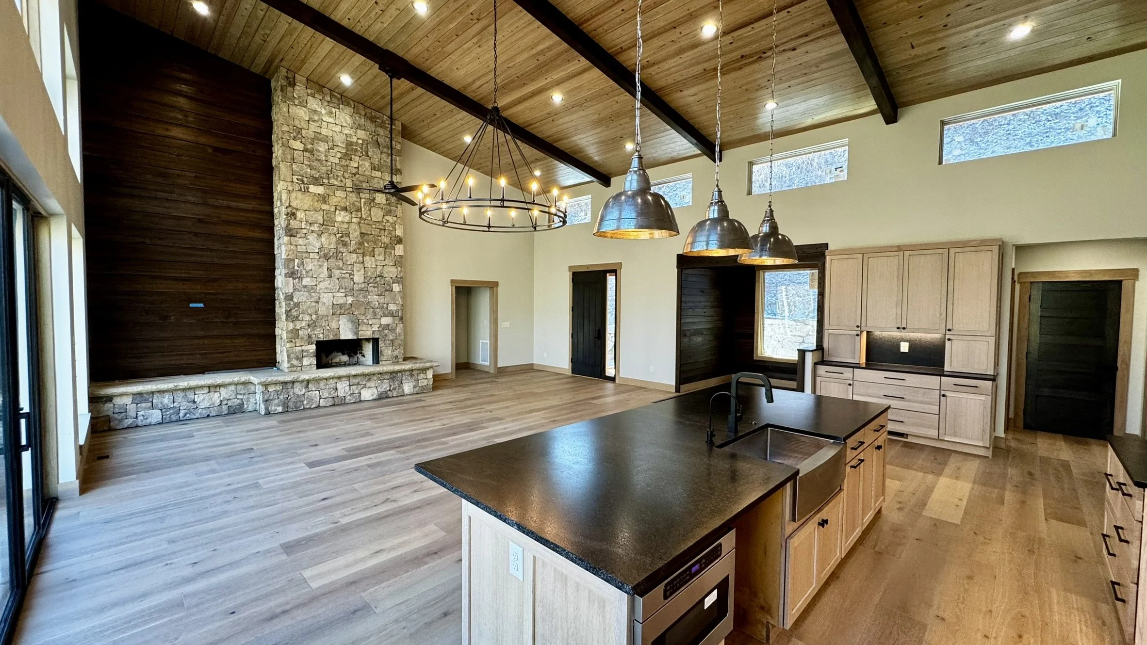 Open-concept living area with a high vaulted wooden ceiling, exposed beams, stone fireplace, large windows, and a kitchen island with a dark countertop and light wood cabinets.