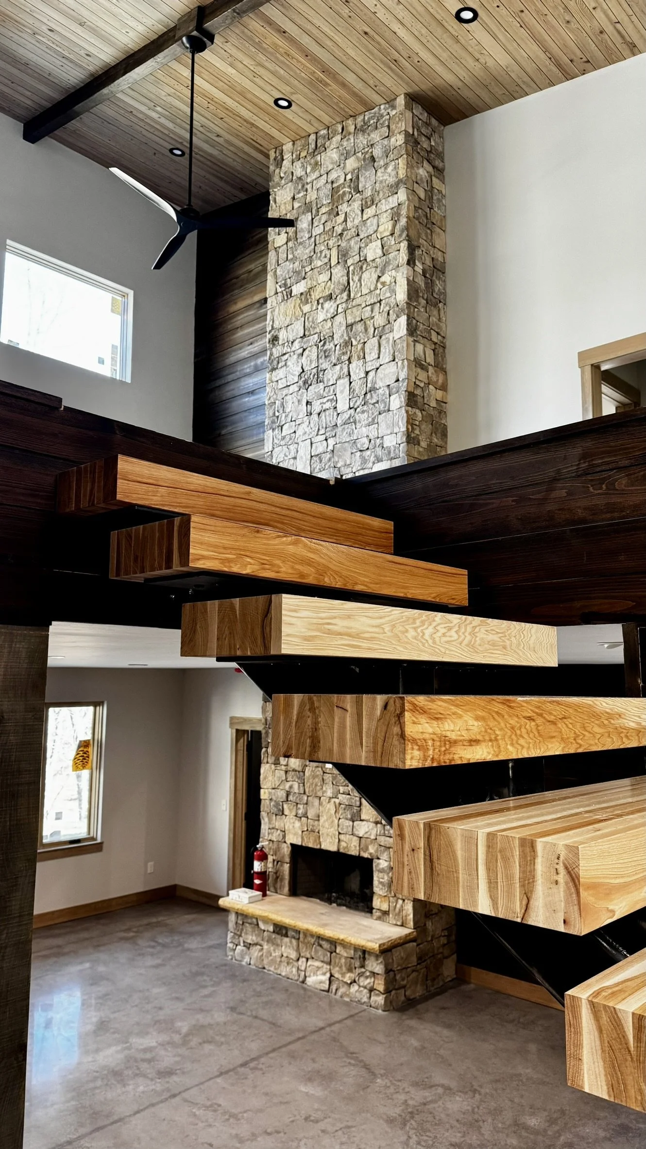 Modern interior with wooden floating stairs, stone fireplace, wood ceiling with recessed lighting, ceiling fan, and windows letting in natural light.