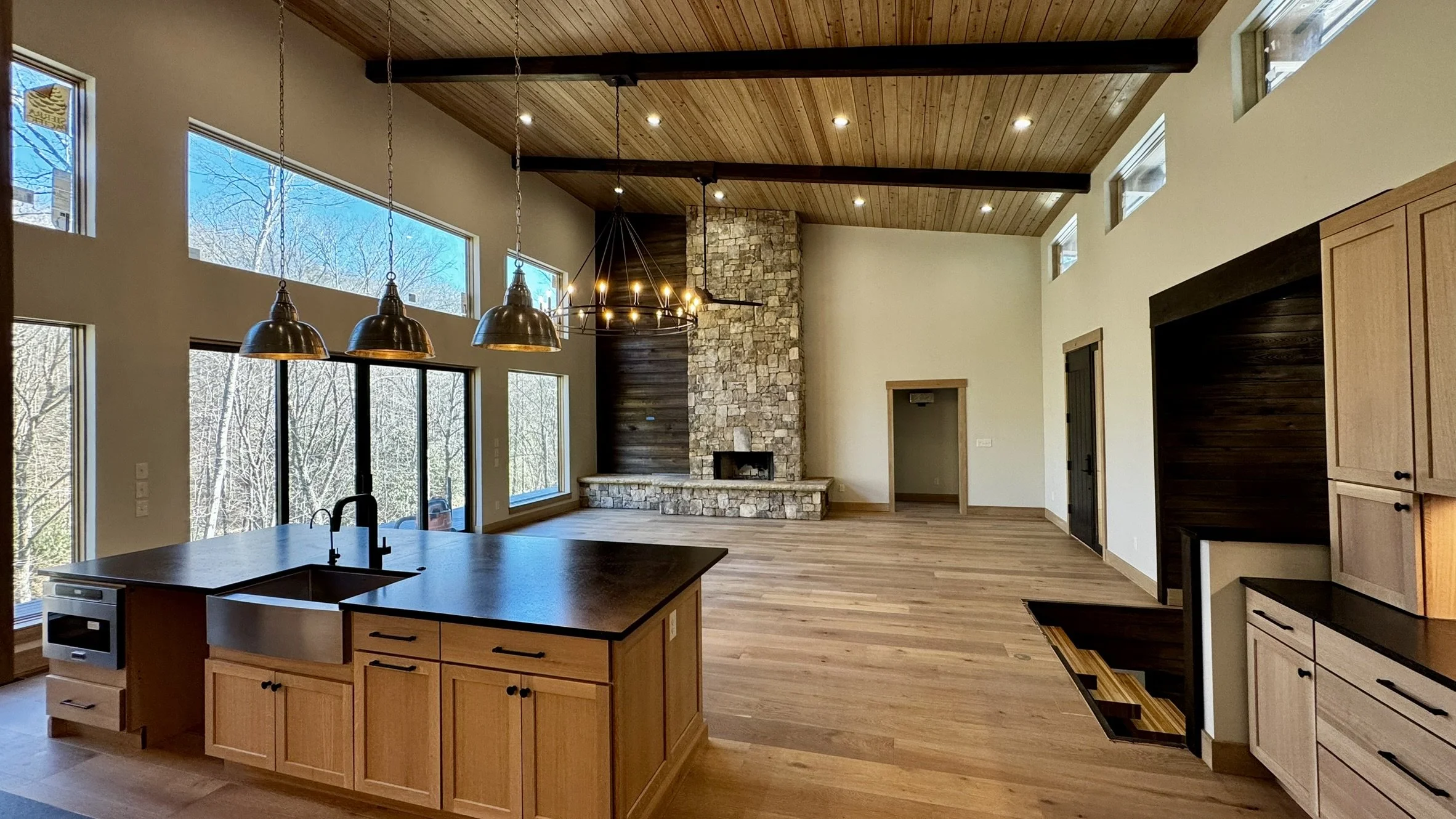 Open-concept kitchen and living room with large windows, wood floors, and a stone fireplace.