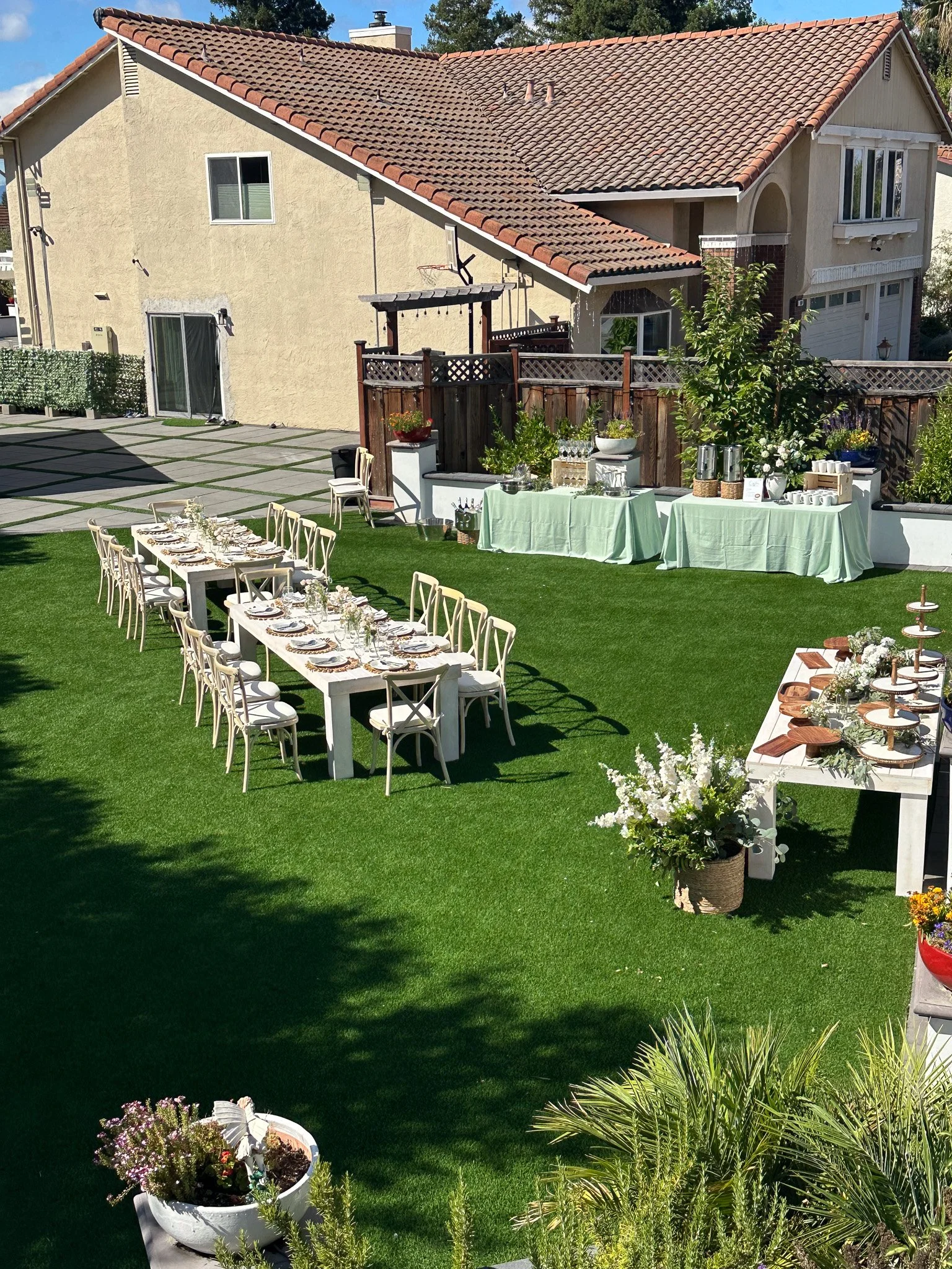Backyard outdoor party setup with long dining tables, white chairs, and tables with floral decorations and serving dishes, on a green lawn with a house in the background.