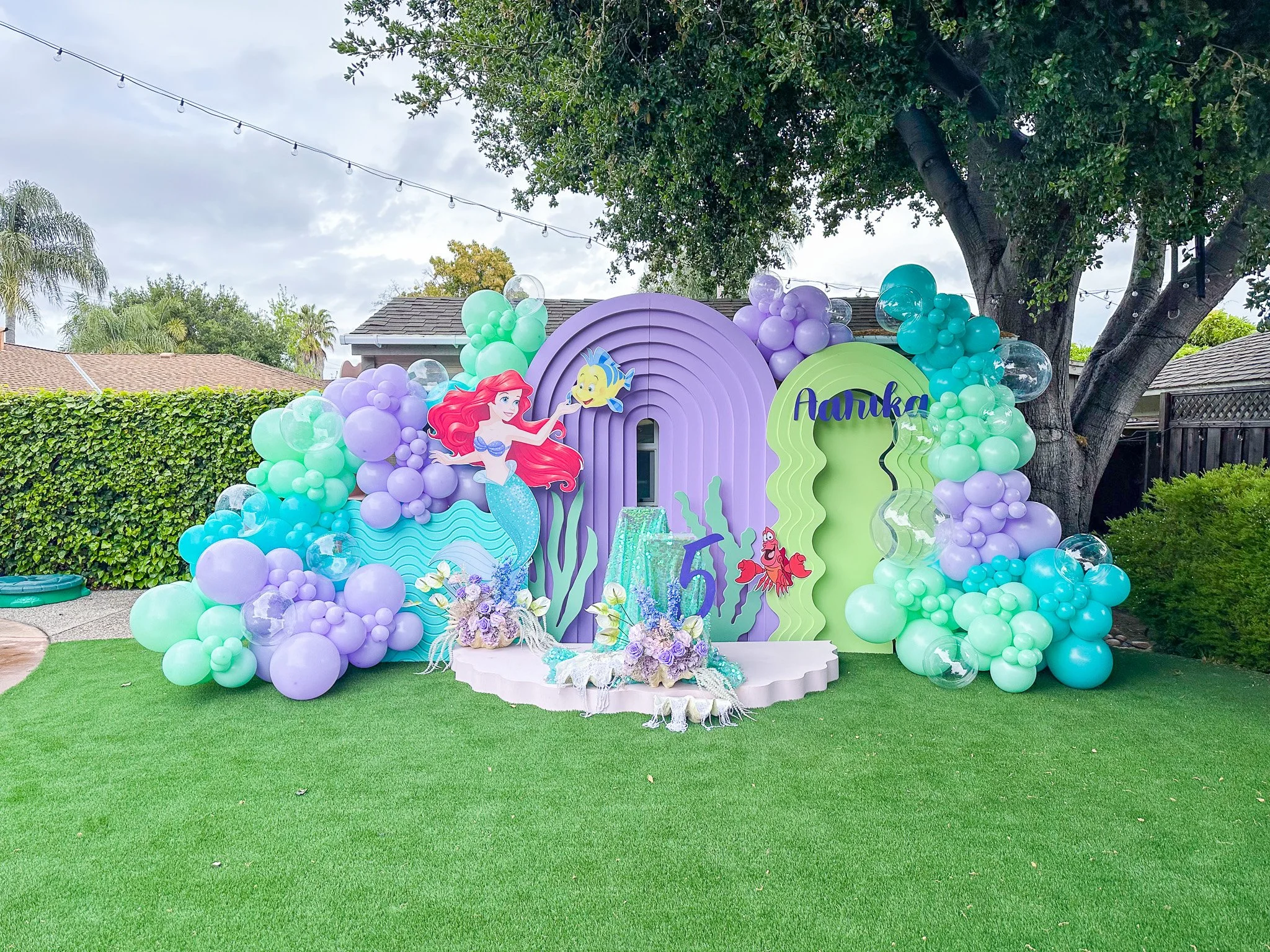 Underwater themed birthday party backdrop with balloons, a large cutout of Ariel from The Little Mermaid, the name Aunika, and a number 5, set on a grassy lawn with trees and houses in the background.