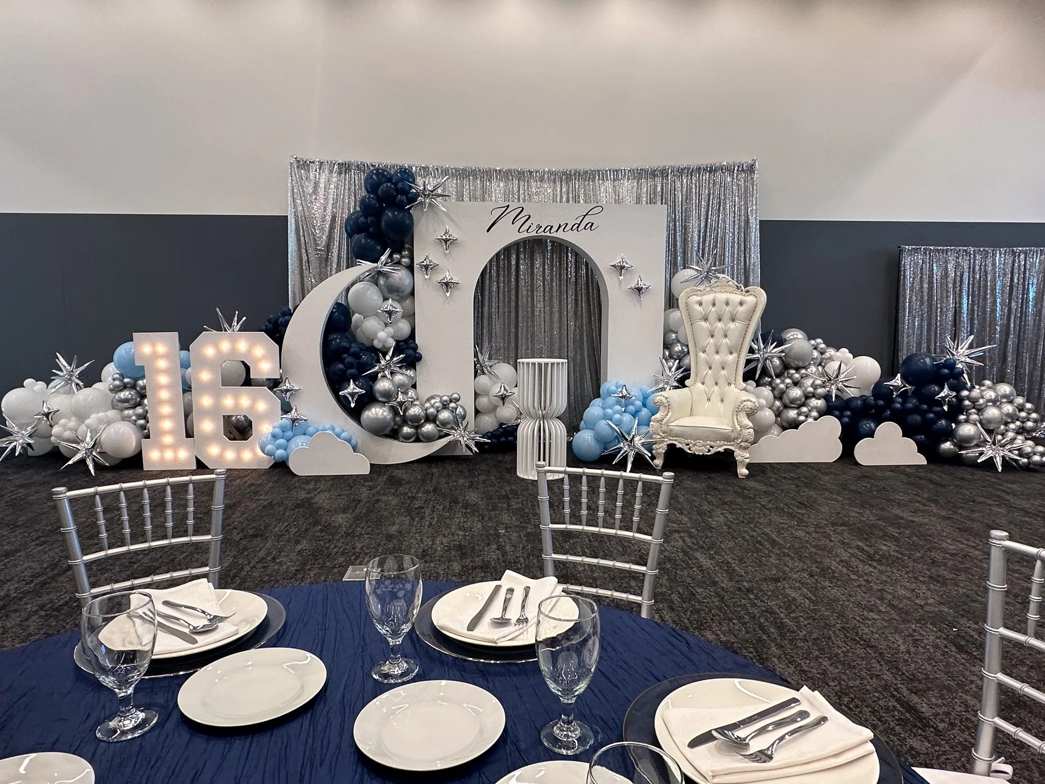 Birthday celebration table with decor for Miranda's 16th birthday, featuring large illuminated number 16, balloon arrangements in white, silver, and navy, star-shaped decorations, a white ornate throne-style chair, and a silver backdrop.