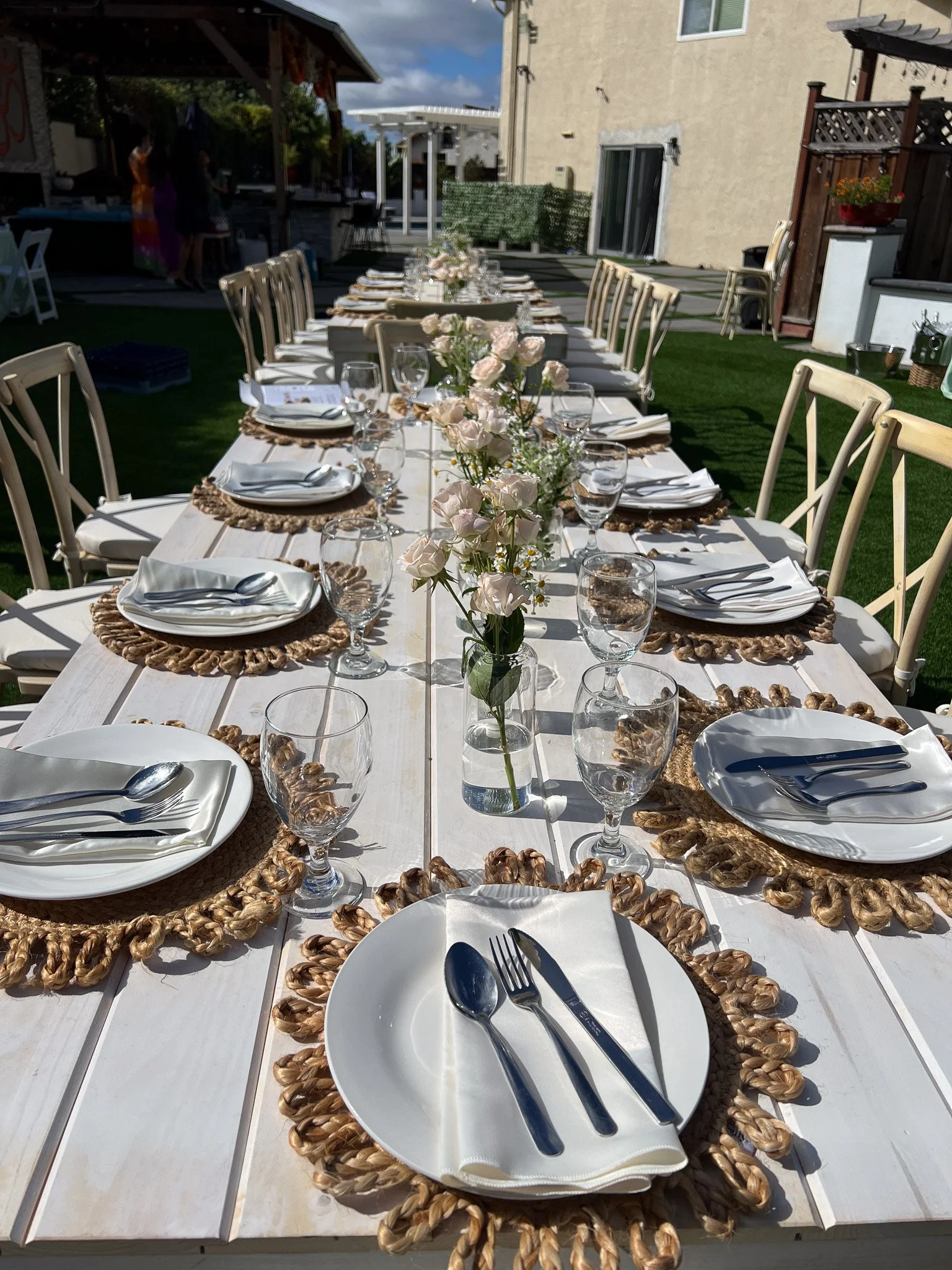 Long outdoor dining table set for a gathering, with white plates, silverware, glasses, floral centerpiece, and woven placemats, in a backyard with green grass and a beige building in the background.