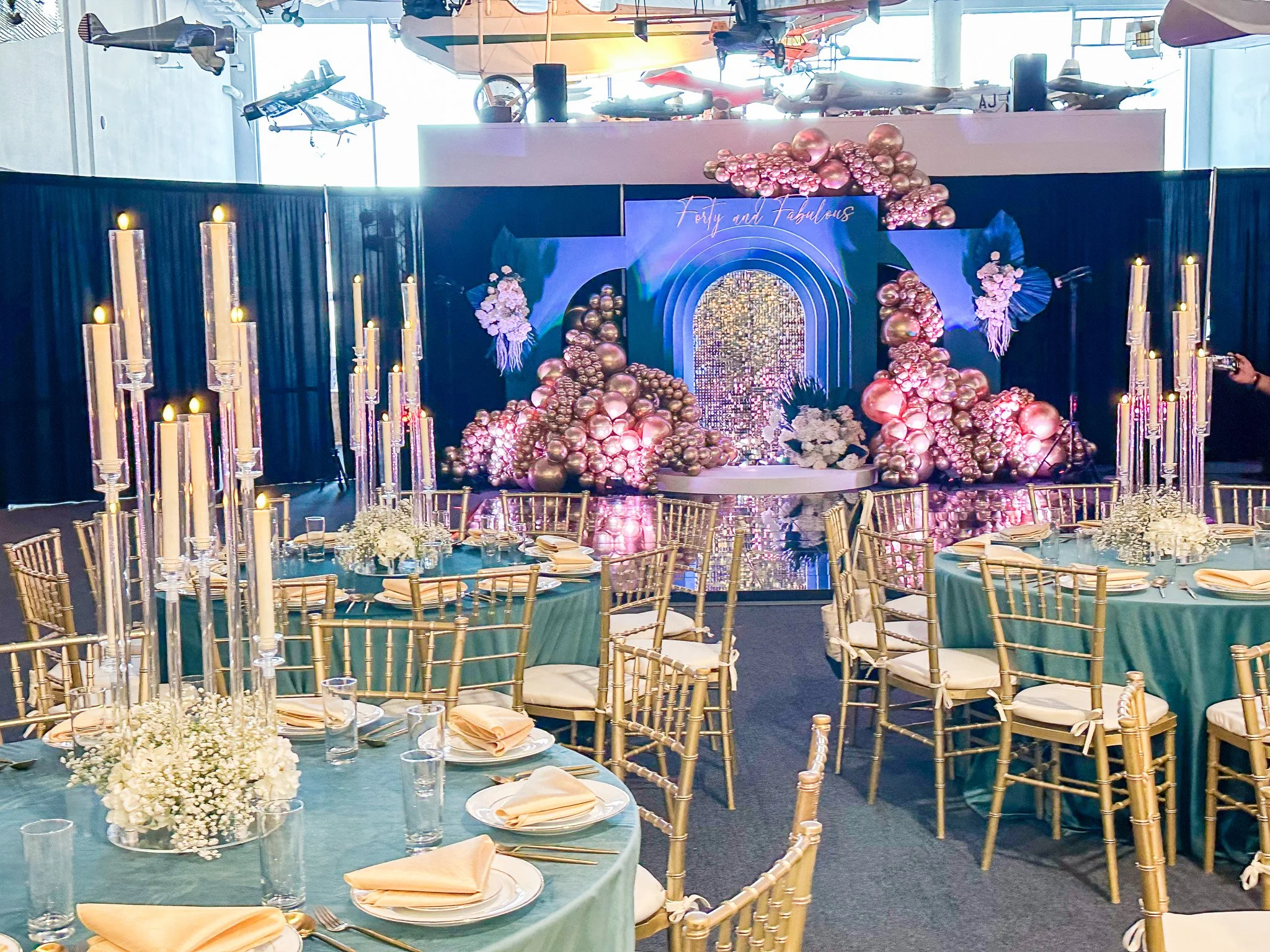 Elegant banquet hall decorated for a celebration with round tables covered in teal tablecloths, gold chairs, and floral centerpieces. The backdrop features a large display of pink and gold balloons, white flowers, and a sign that reads 'Fifty and Fabulous', indicating a 50th birthday celebration. Tall candles in glass holders are on the tables and ceiling-mounted model airplanes are visible.