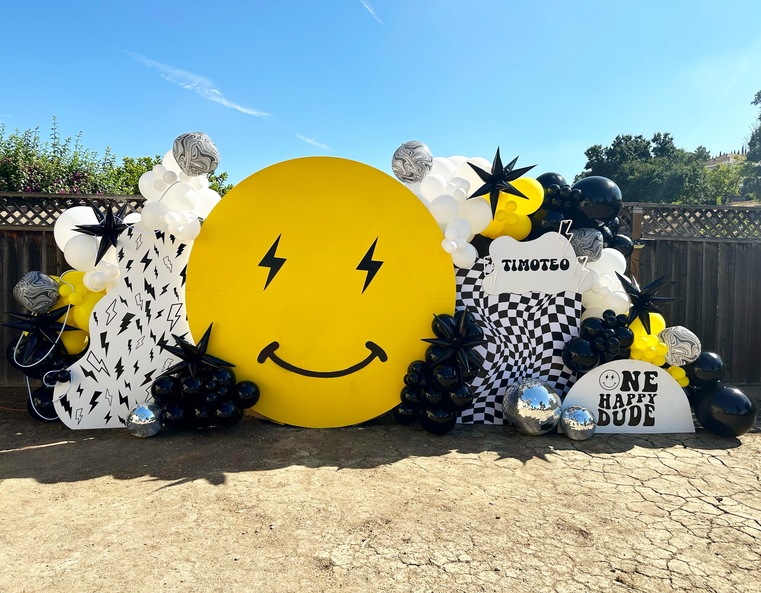 Decorative outdoor setup with a large yellow smiley face, black lightning bolt eyes, surrounded by black, white, and yellow balloons, checkered and lightning bolt-themed banners, and signs reading 'Timoteo' and 'One Happy Dude'.