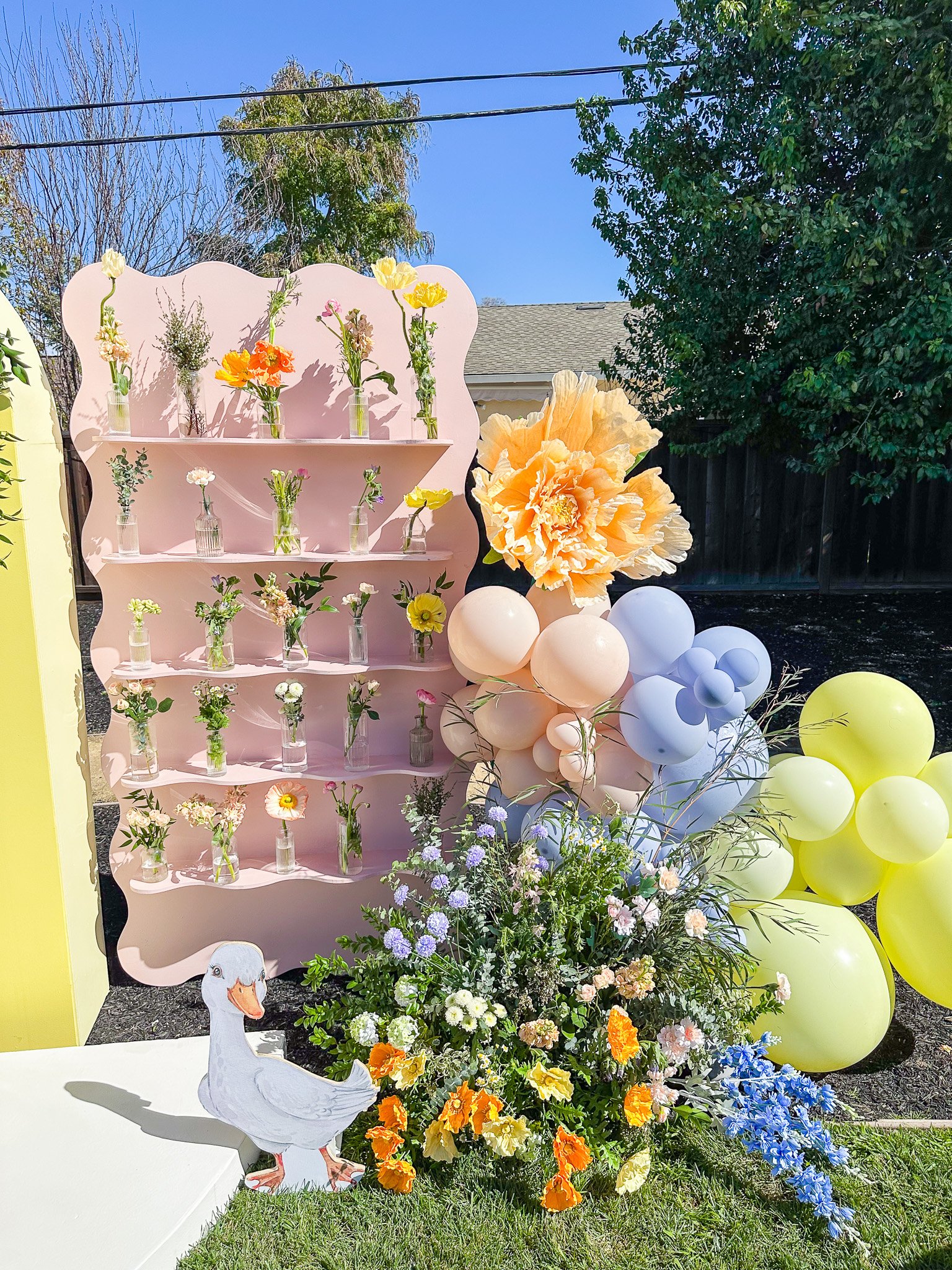 Decorative outdoor setup with a pink shelf displaying small vases of flowers, surrounded by colorful balloons in pastel shades and a large paper flower. There is a large flower and a cardboard goose cutout in front, with a vibrant flower arrangement growing at the base, and trees and a house in the background.