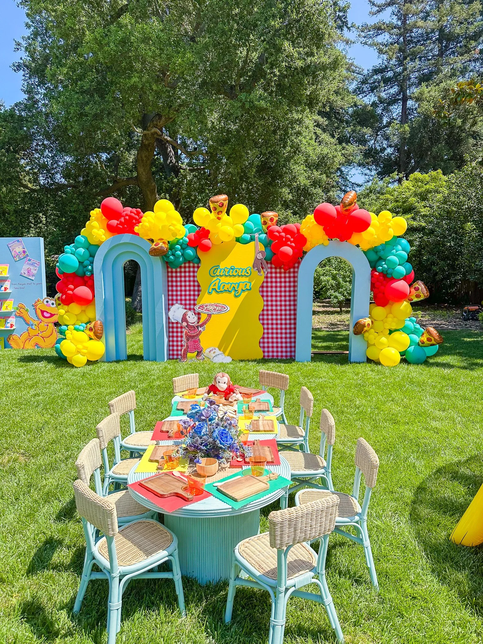 Colorful outdoor birthday party setup featuring a balloon arch with red, yellow, and turquoise balloons, and a backdrop with a yellow sign reading 'Curious Aarya' decorated with cartoon characters. There is a round table with colorful placemats, a flower centerpiece, and chairs around it, all set on a grassy lawn with tall trees in the background.