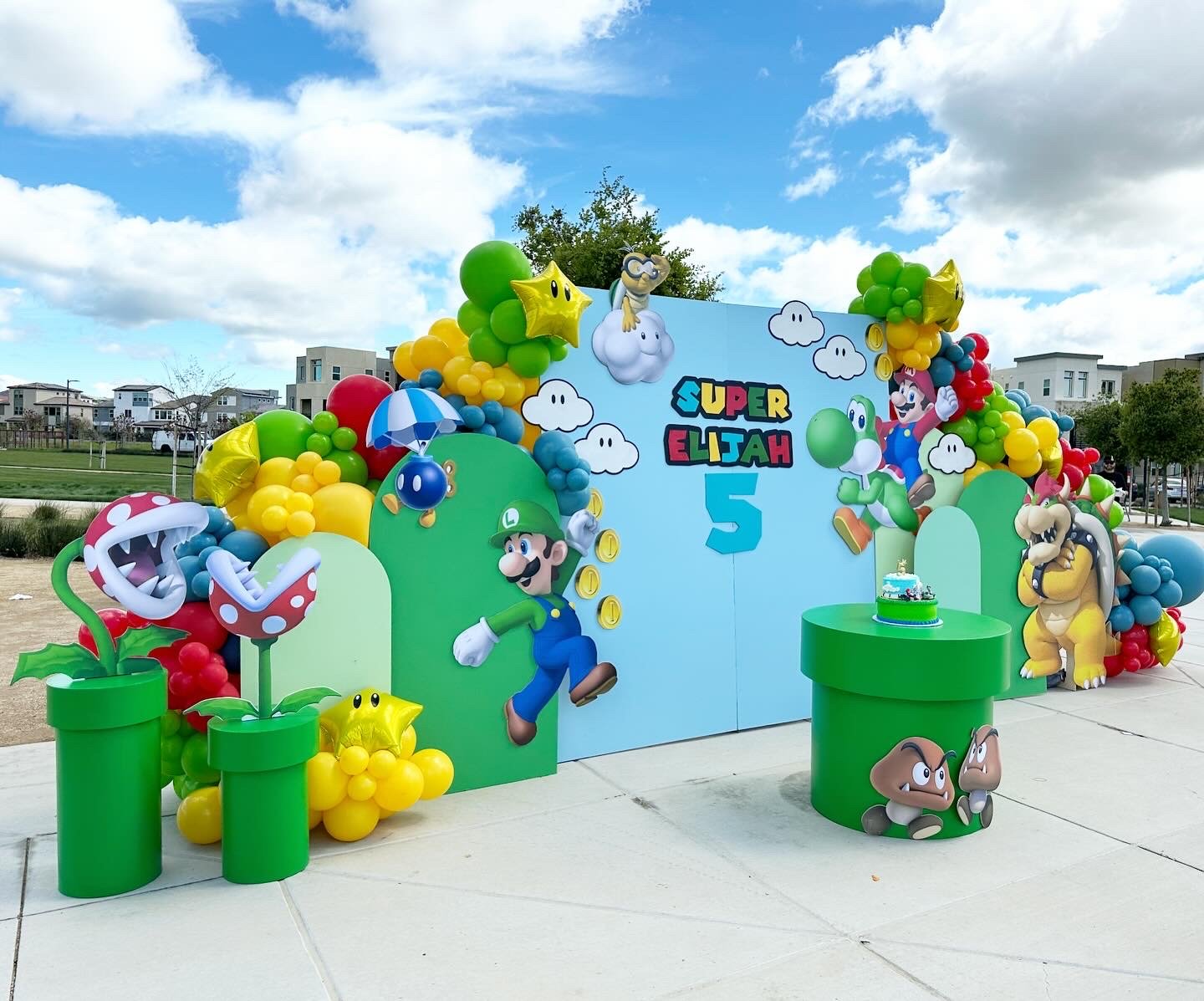 Super Mario-themed birthday party setup with colorful balloons and character cutouts, featuring Luigi, Mario, Bowser, and other Mario characters, with a backdrop displaying the name 'Super Elijah' and the number 5, and a birthday cake on a green table.