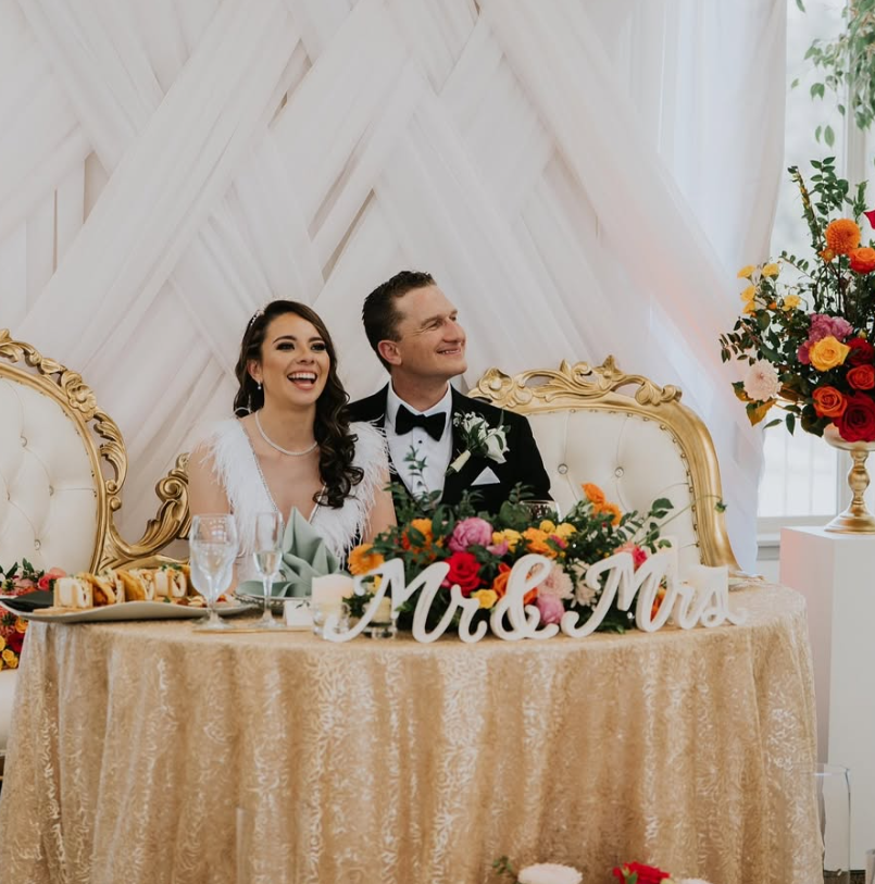 Bride and groom sitting at a wedding reception table, smiling, with a gold and cream-colored backdrop, floral arrangements, and wedding signs that say 'Mr. & Mrs.'