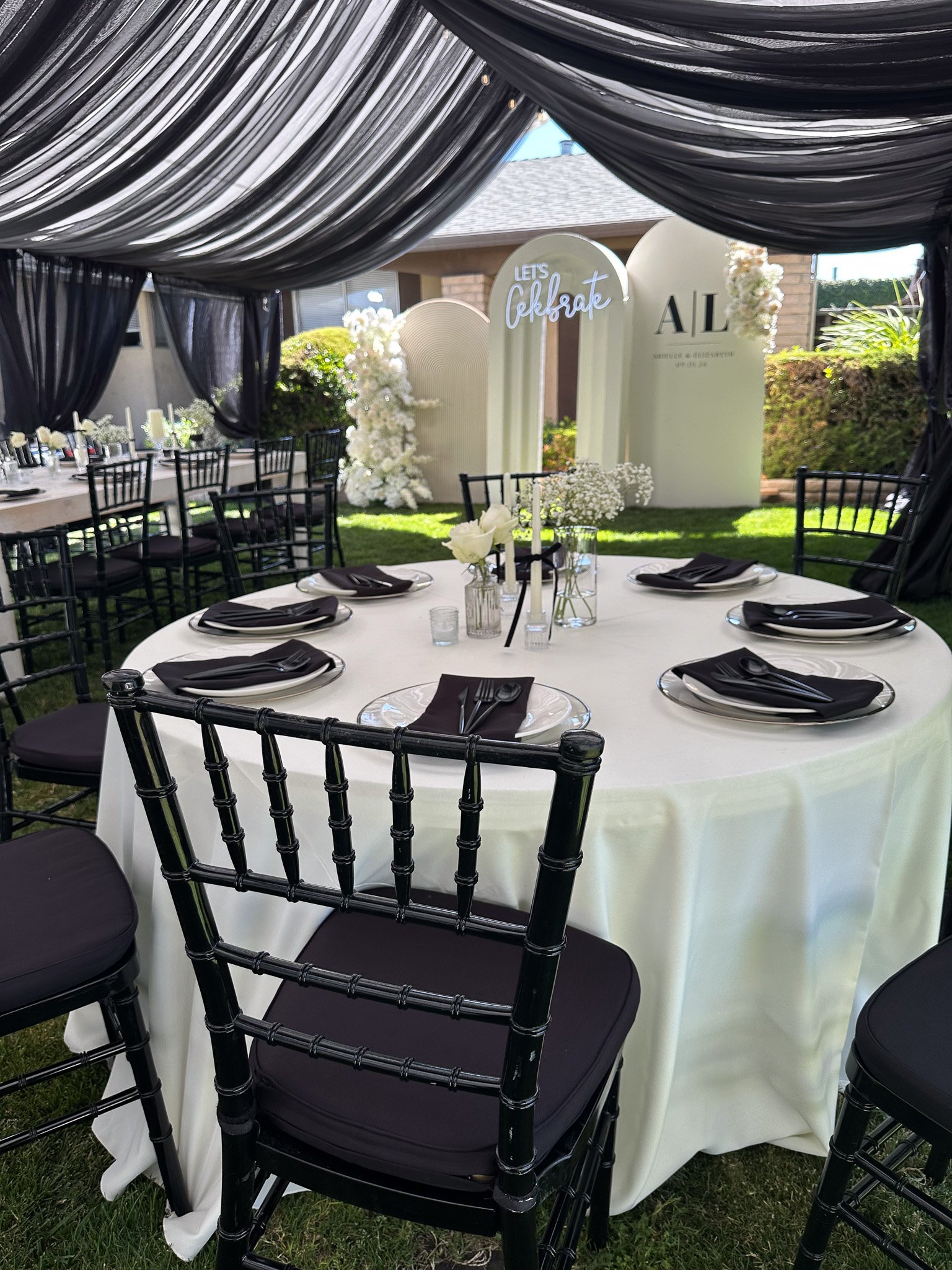 A round table set for an outdoor celebration under a black and white draped canopy, with black chairs, floral centerpieces, and a backdrop with the words "Let's Celebrate" and "AL" visible.