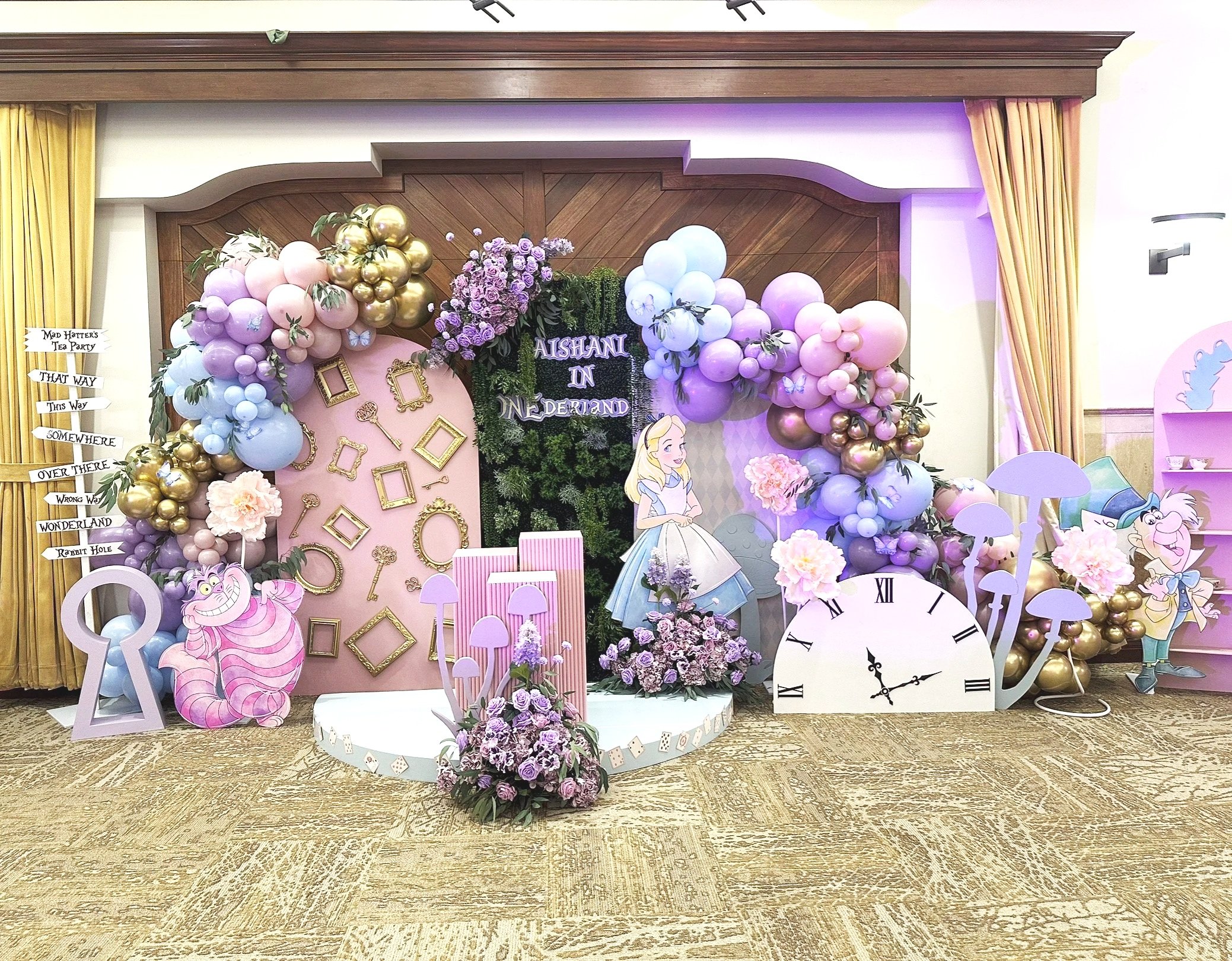 A decorated backdrop featuring characters from Alice in Wonderland, with balloons, flowers, frames, and themed props in pastel and gold colors, set for a party or event.