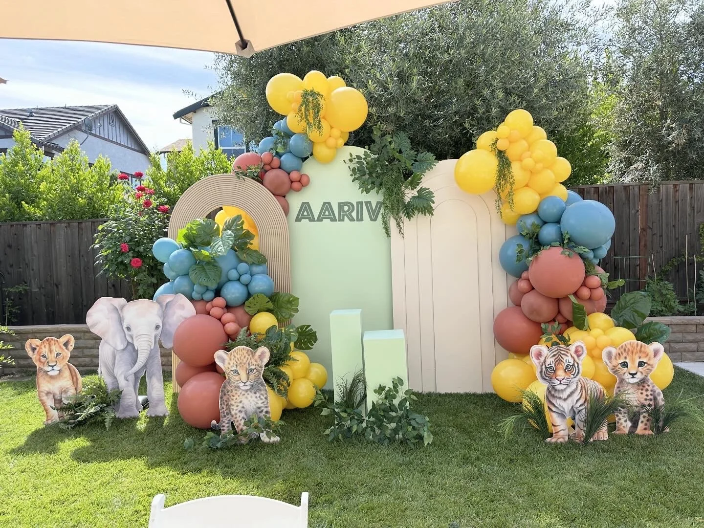 A jungle-themed birthday party backdrop with balloons arranged around wooden panels and greenery, featuring cutouts of various baby jungle animals including a lion cub, elephant, leopard, tiger cub, and cheetah cub. The name 'AARIV' is displayed at the top of the backdrop.