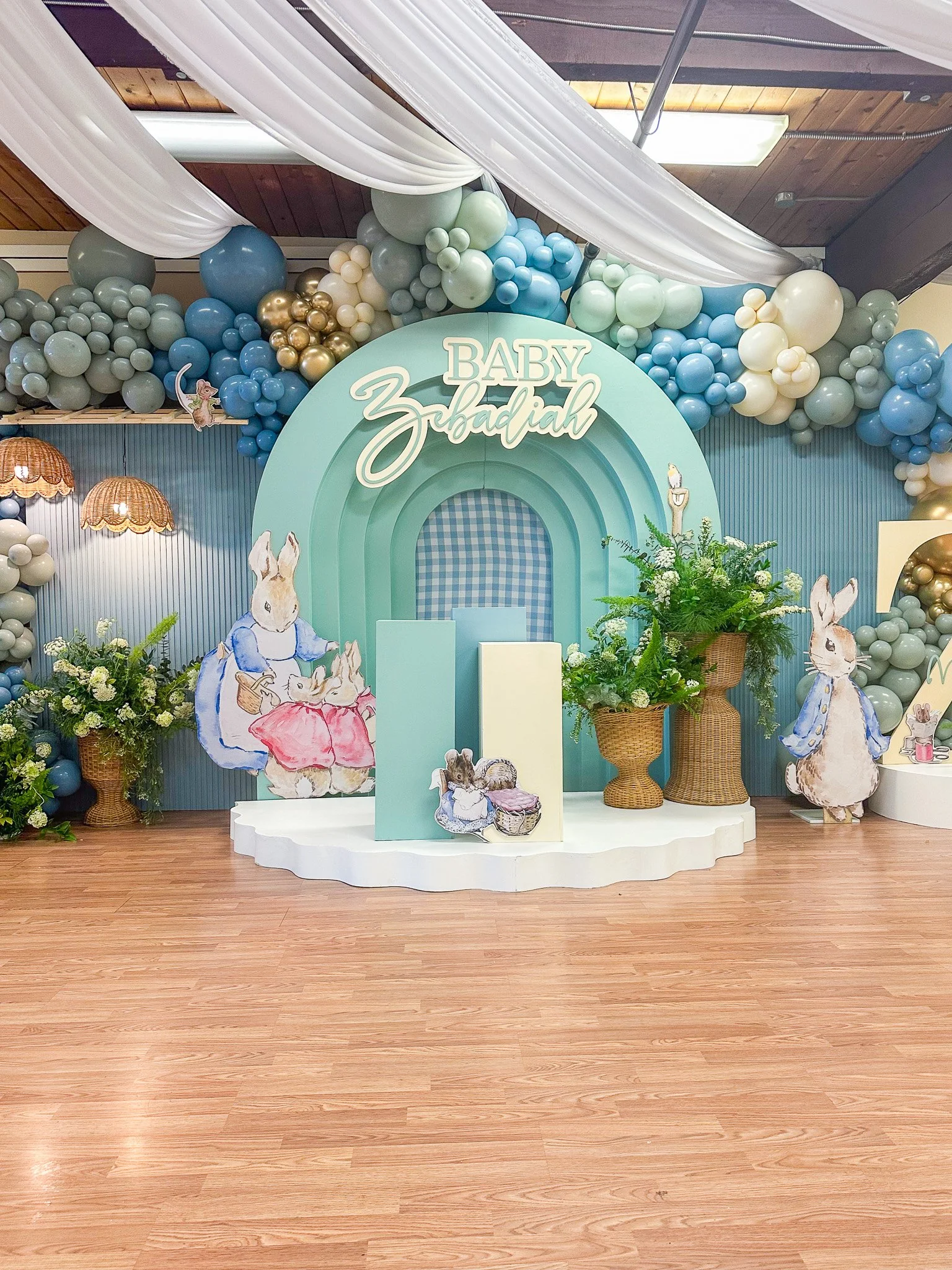 Decorative setup for a baby's shower or celebration featuring a pastel blue and white balloon arch, illustrated bunny characters, floral arrangements, and a backdrop with the words "Baby Zebadiah".