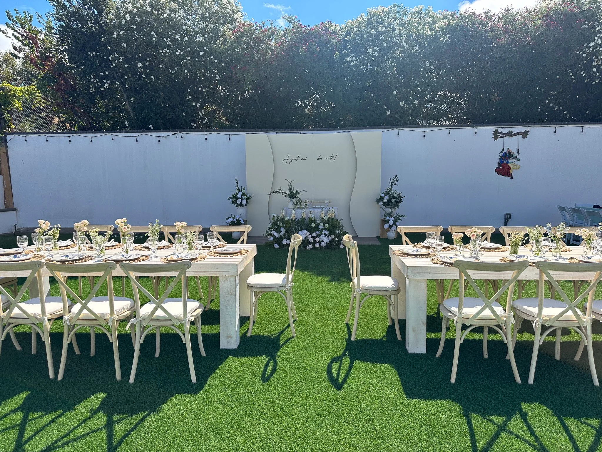 Outdoor wedding reception area with white tables and chairs, decorated with floral centerpieces, set on a green lawn with a backdrop of floral arrangements and a white wall with a decorative sign.