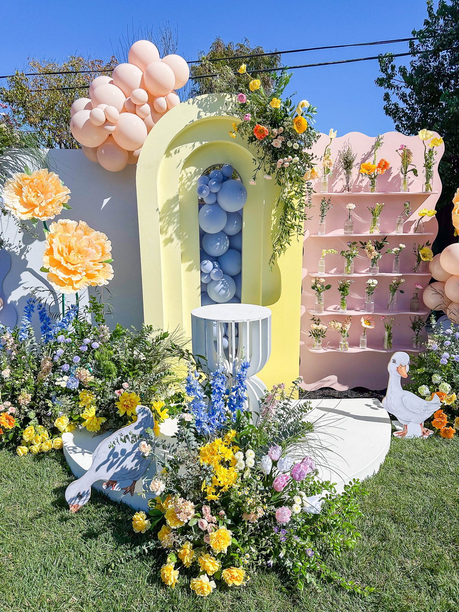 Decorative outdoor display with pastel-colored balloons, arranged flowers in vases, cutout ducks, and a floral backdrop against a blue sky.
