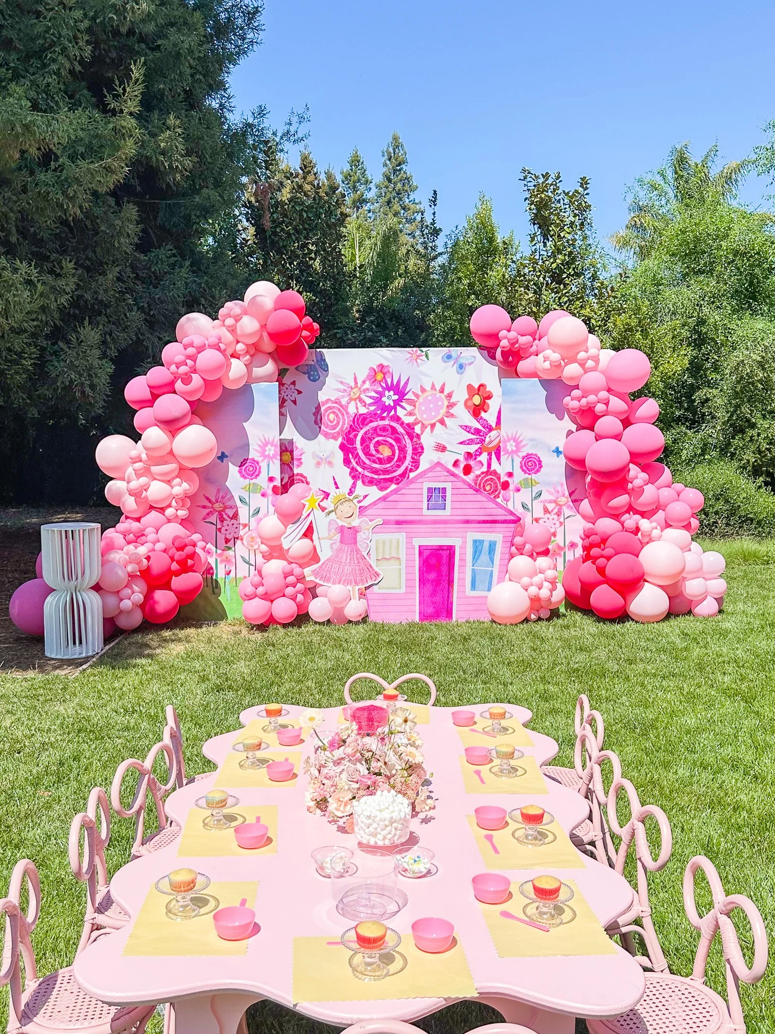 Outdoor birthday party setup with pink theme, featuring a decorated backdrop with balloons and a house illustration, a pink table with chairs, and a floral centerpiece, surrounded by trees under a blue sky.