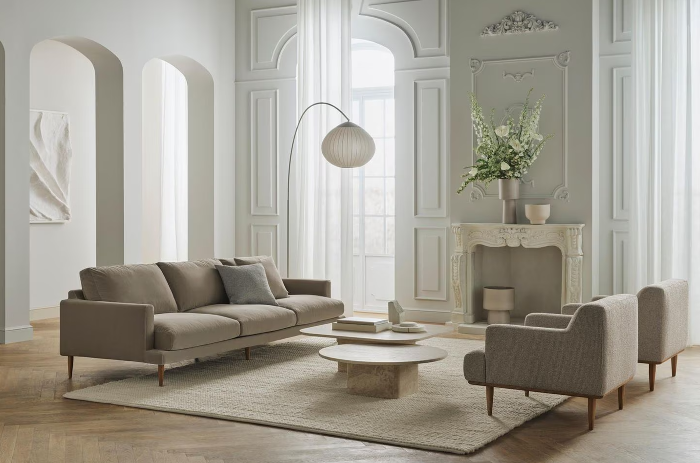 Sala de estar con sofá beige, dos sillones grises, mesas de centro de piedra, chimenea decorativa con florero y flores blancas, cortinas blancas, pared decorativa y lámpara de pie