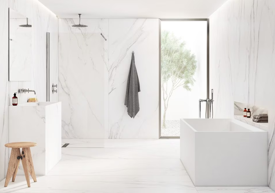 Baño moderno con paredes y piso de mármol, ducha con doble cabezal, bañera rectangular, ventana grande con vista a un árbol, estantería con champús, toallas y productos de cuidado personal, taburete de madera, y una toalla gris colgada.