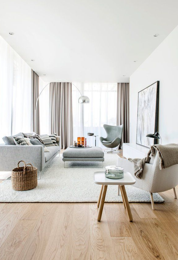 Sala de estar moderna con sofá gris, sillón de oreja gris, lámpara de arco, cortinas claras, alfombra beige y mesa de centro con decoración.
