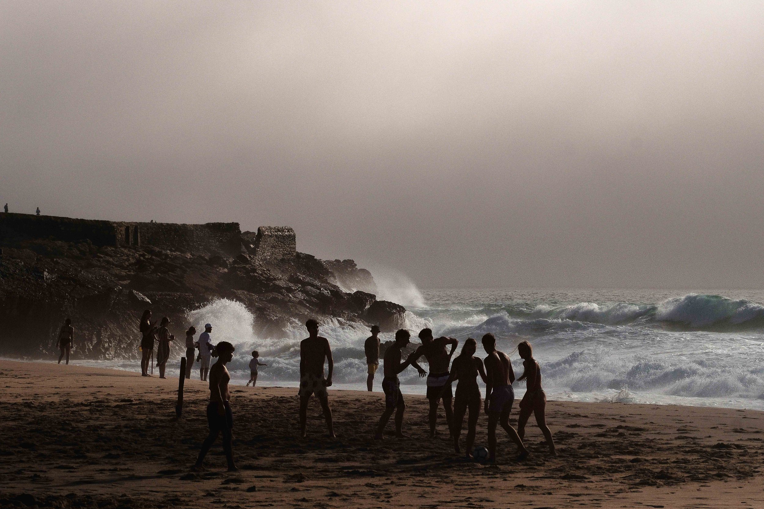People on a beach by the ocean, with waves crashing against rocks.