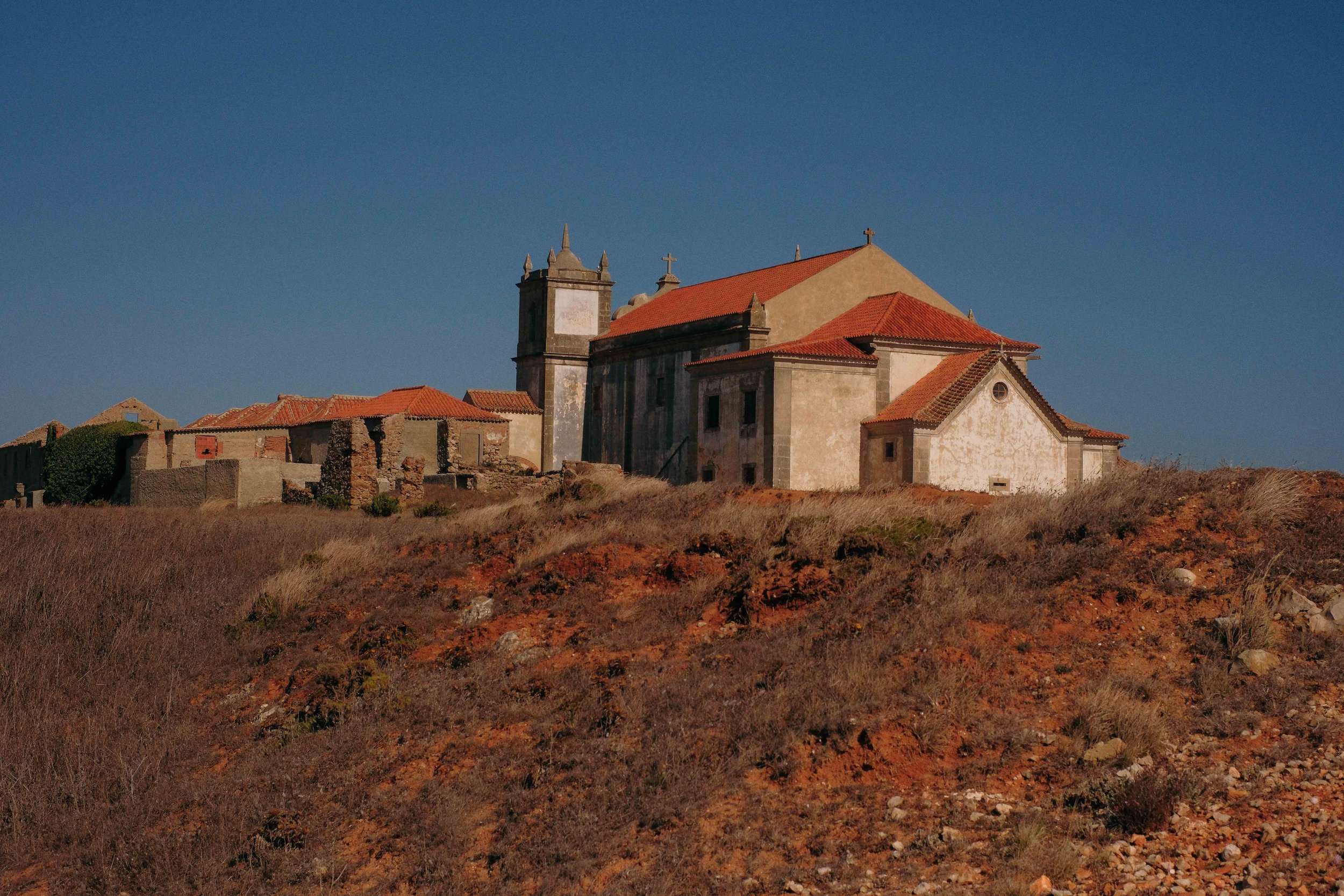Old church with red-tiled roof on a dry hillside under a clear blue sky, surrounded by ruins.