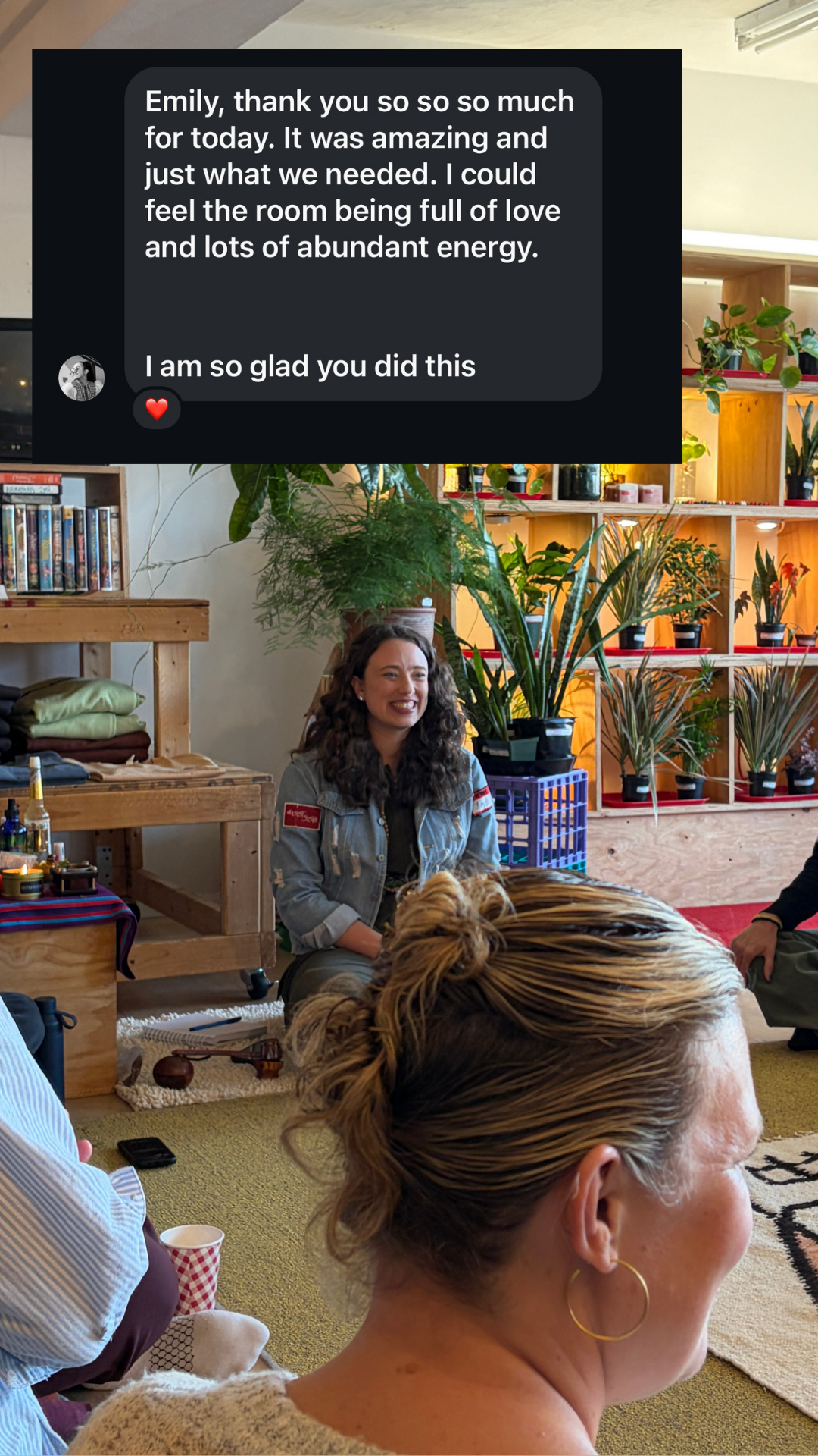 A group of people sitting on the floor in a cozy indoor setting, with a woman with curly hair smiling and wearing a denim jacket. There are plants and wooden shelves in the background, along with a person with blonde hair tied in a bun, and some casual items like a cup and a remote on the floor.