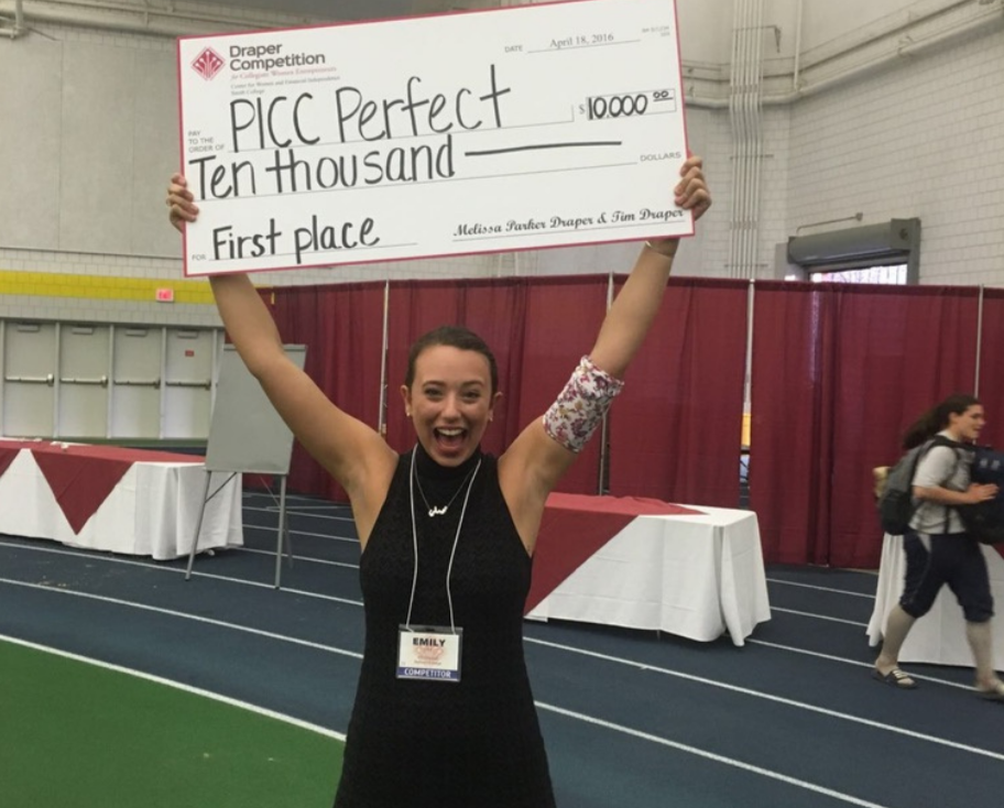 A young woman with a big smile, wearing a black sleeveless dress and a name tag that says Emily, holding up a giant check for $10,000 for first place at a competition, inside a gymnasium with banners and red curtains in the background.