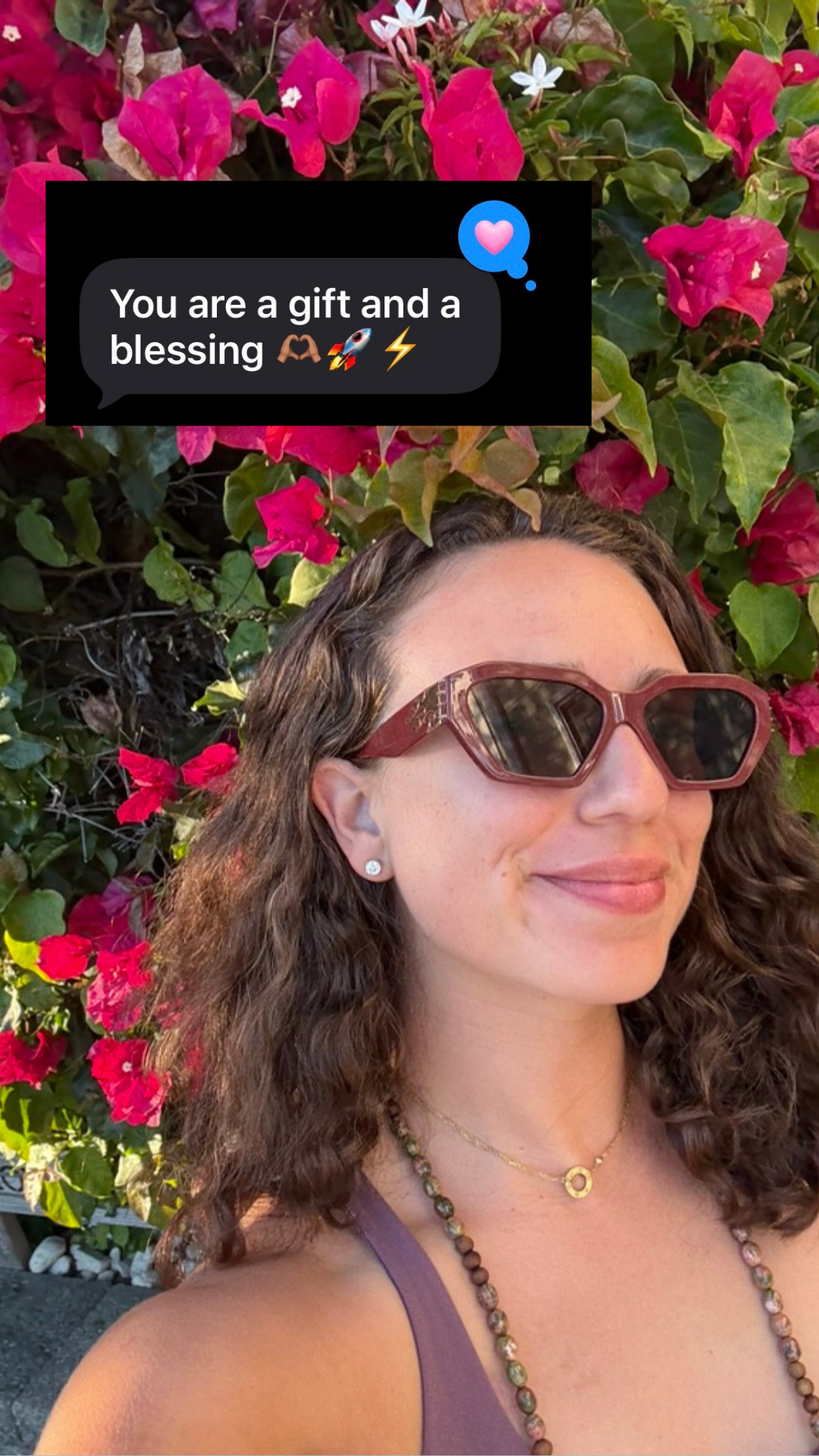 A woman with curly brown hair wearing sunglasses and jewelry, standing in front of pink and red flowers, smiling.