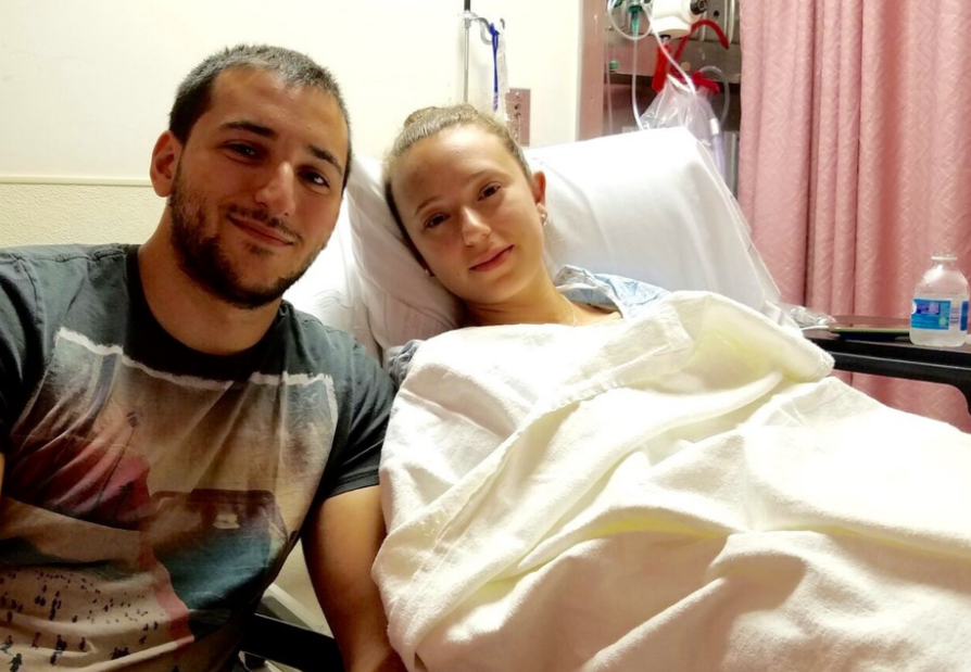A young woman lying in a hospital bed, hospitalized and covered with a blanket, with a man taking a selfie next to her, both smiling.