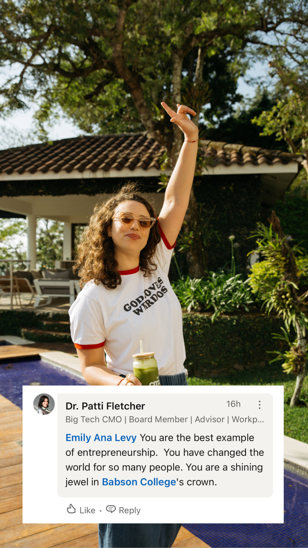Woman with curly hair and sunglasses holding a green smoothie, standing outdoors near a pool and trees, wearing a white T-shirt with red trim that reads 'GOD LOVES WEIRDOS'.