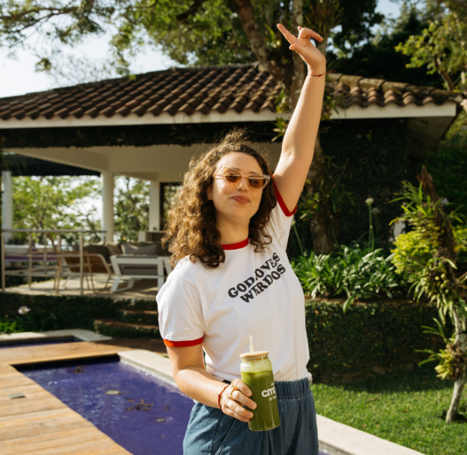 Woman with curly hair wearing sunglasses and a white T-shirt with red trim, holding a green smoothie, raising her arm and smiling outdoors near a pool and garden.