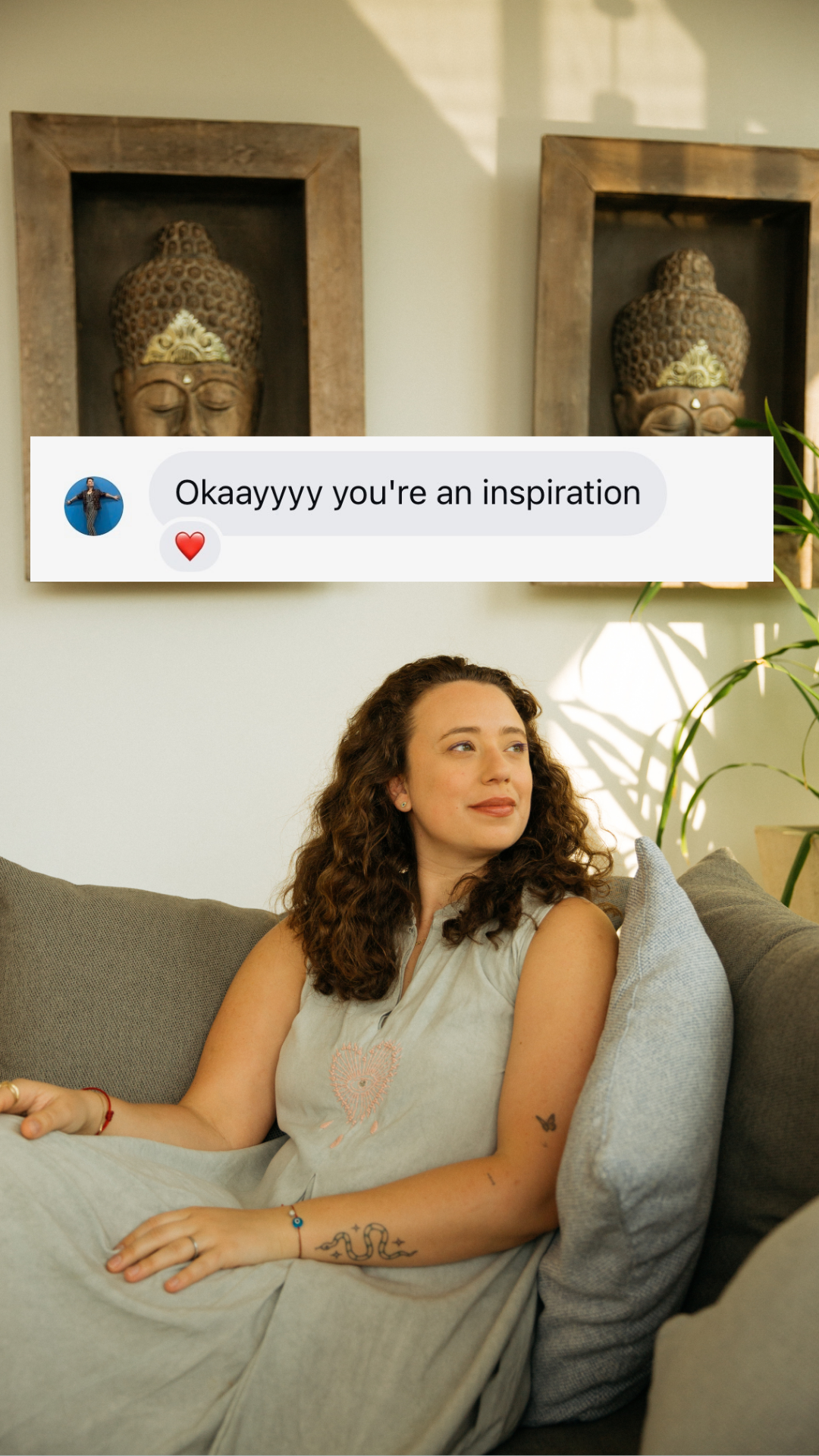 A woman with curly brown hair sitting on a gray couch, looking to her left with a slight smile, in a living room with art pieces on the wall and a plant in the background. A text overlay says, 'Okaayyy you're an inspiration' with a heart emoji.
