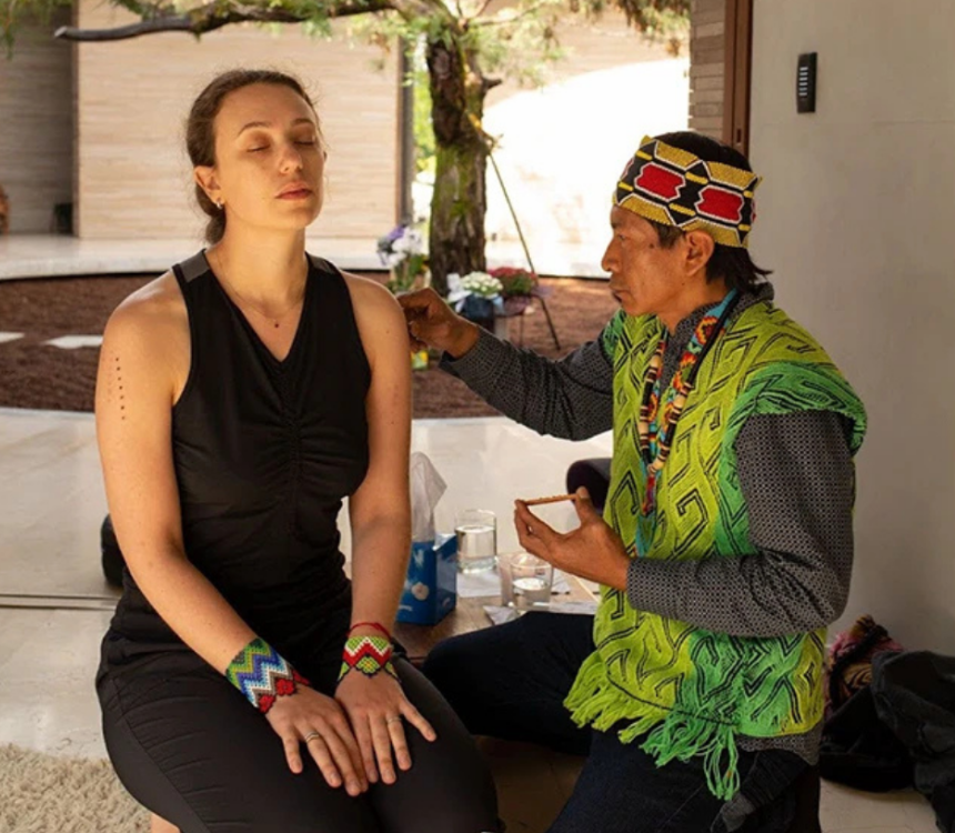 A woman is receiving a traditional indigenous hair and face painting from an elder woman wearing traditional attire outdoors.