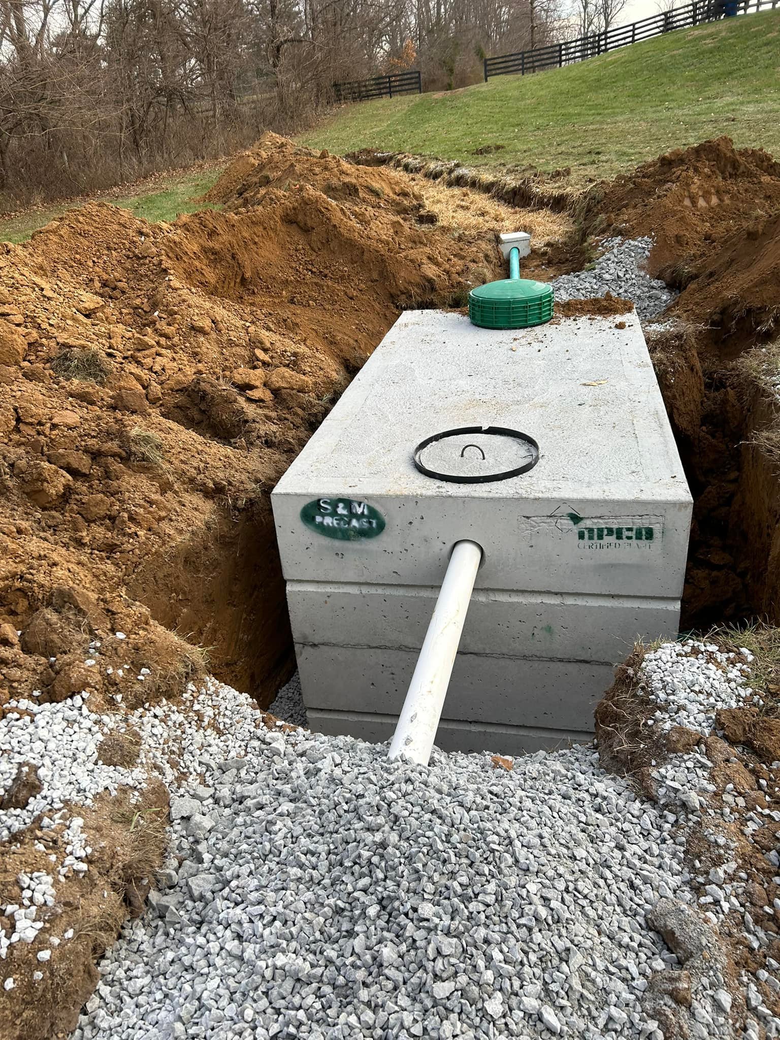 Installation of an underground concrete septic tank with attached pipes, surrounded by gravel and dug earth, in a yard with grass and trees.
